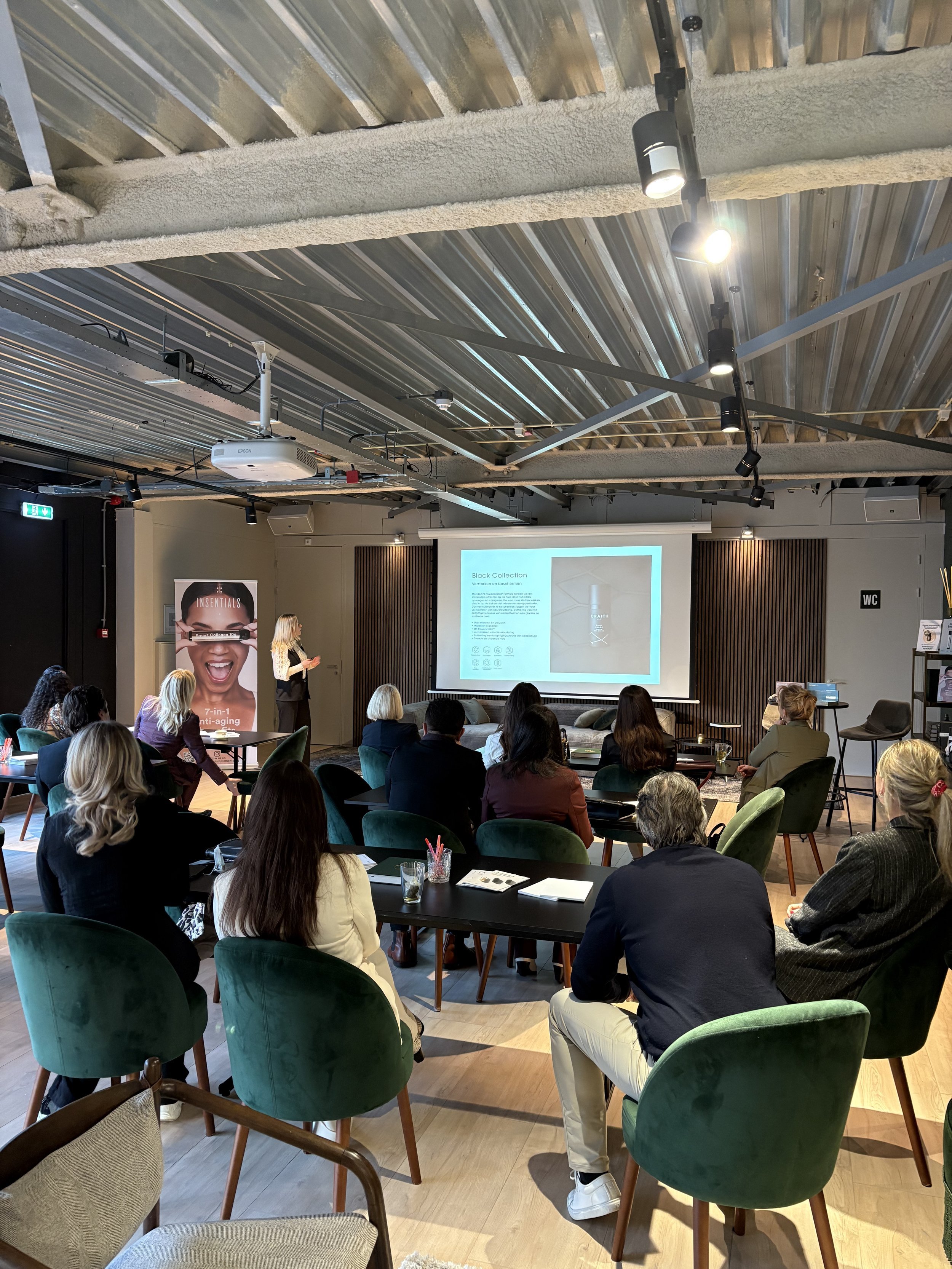 Presentation in a modern room with people seated in green chairs, watching a woman presenting slides on a projector screen.