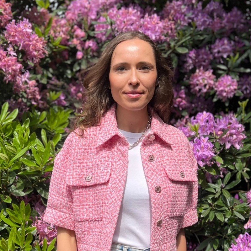 Woman in a pink jacket standing in front of vibrant pink and purple flowers.