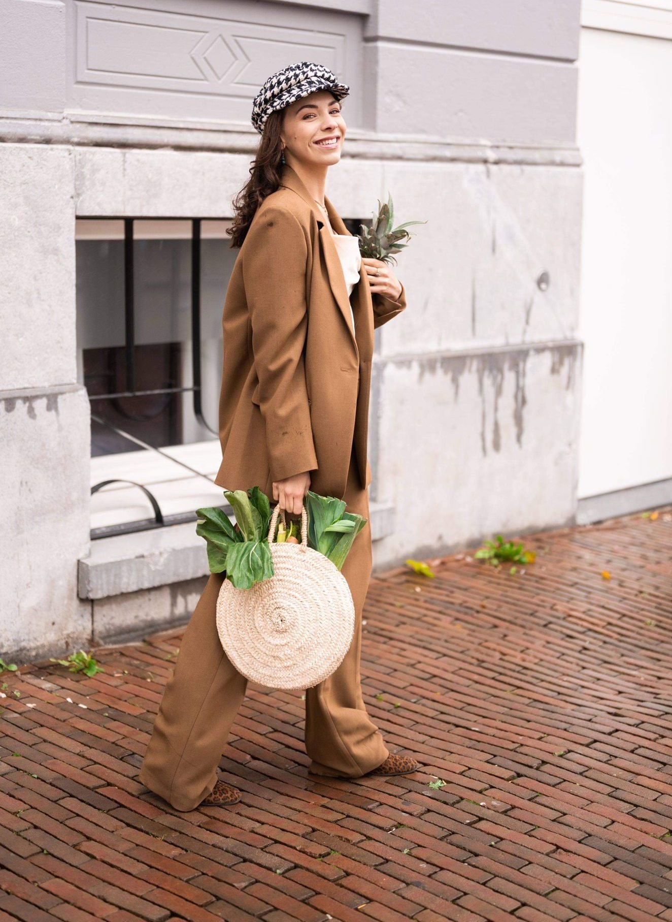 Smiling woman in brown suit and hat holding a round straw bag with green vegetables walking on a brick street.