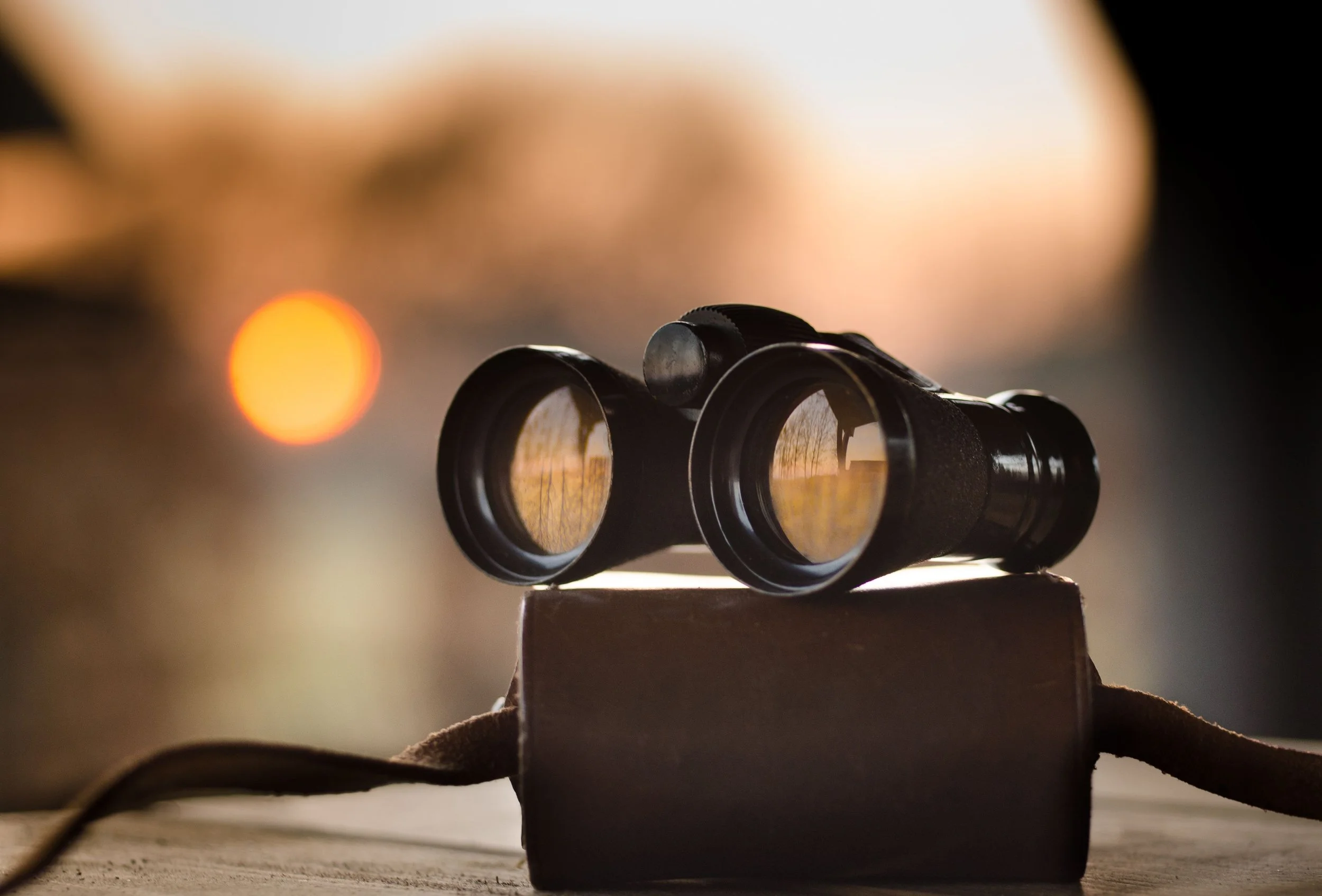 Ein Fernglas liegt auf einem Holztisch, im Hintergrund sind verschwommene Lichter, die einen Sonnenuntergang andeuten.