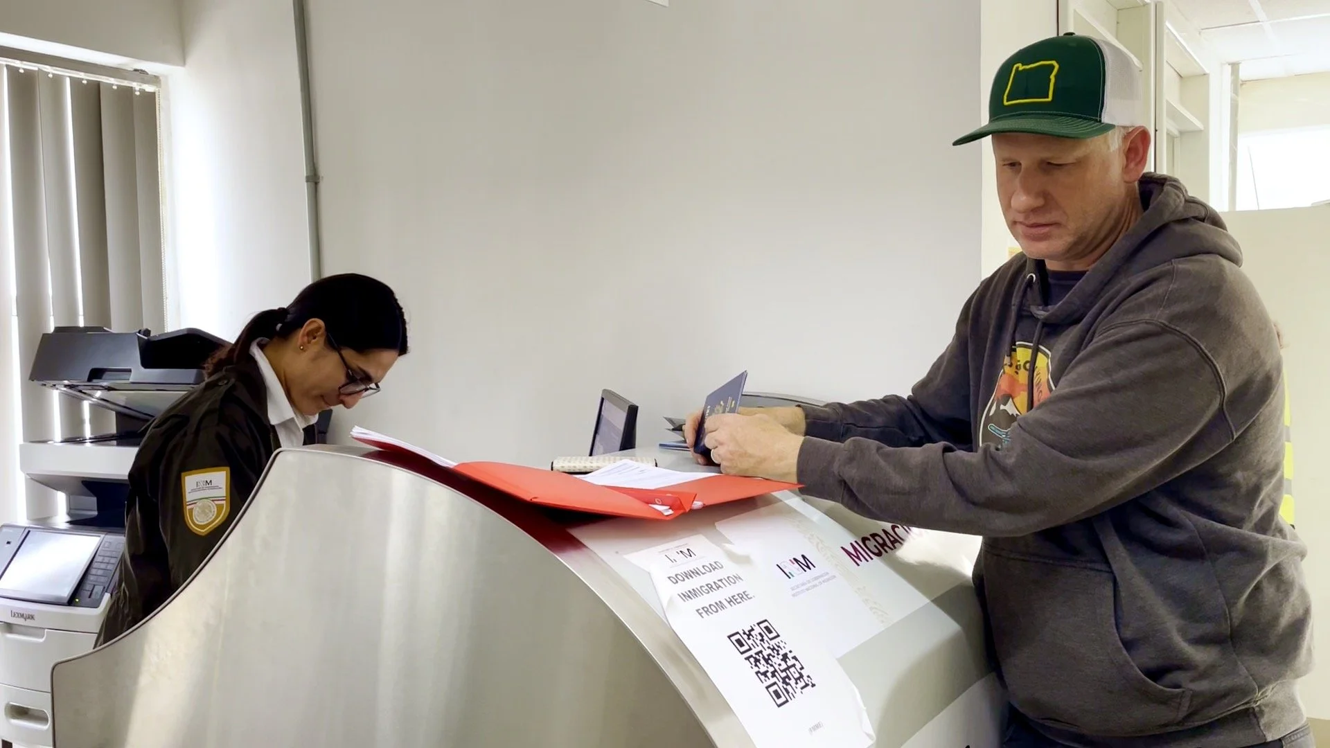 A man at a counter with paperwork and a woman behind it, in an office or service area setting.