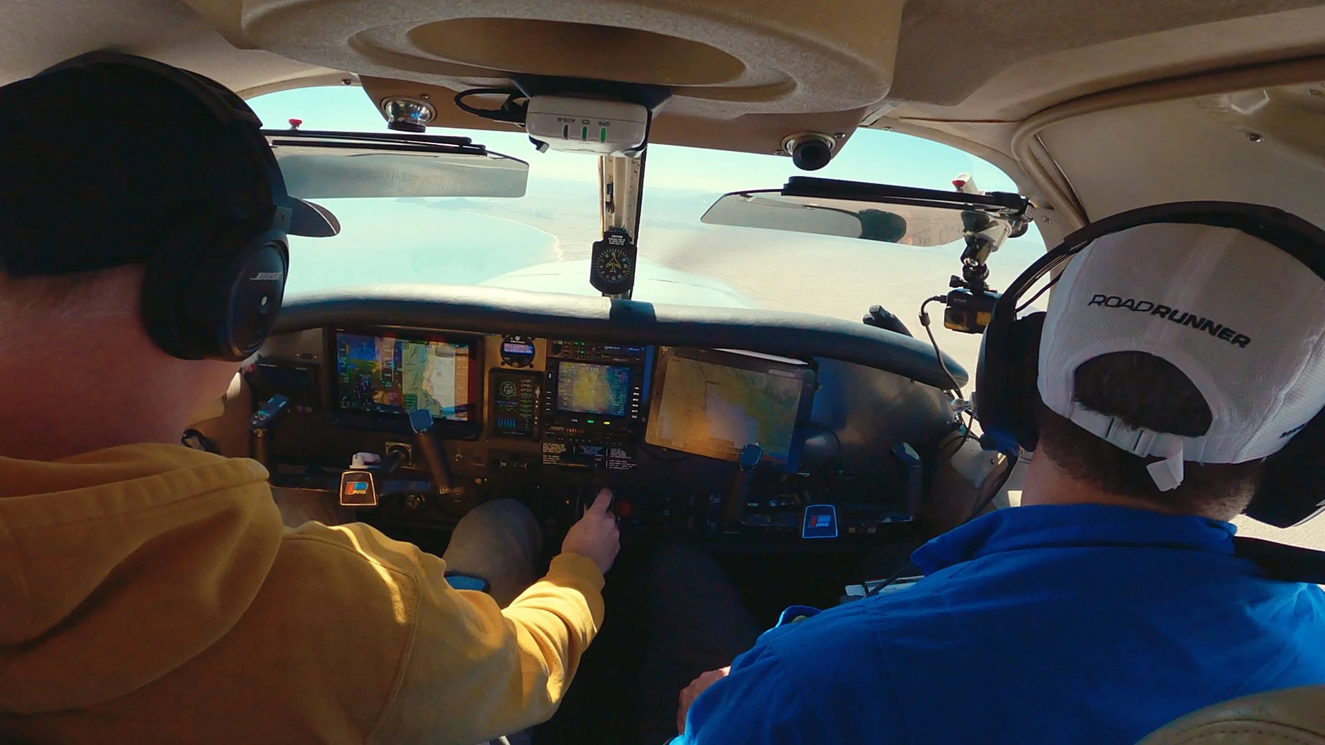 Inside the cockpit of a small airplane with two pilots, one wearing a yellow hoodie and the other a blue jacket, both wearing headsets. The cockpit has multiple screens and controls, and the view outside shows a coastline and water.