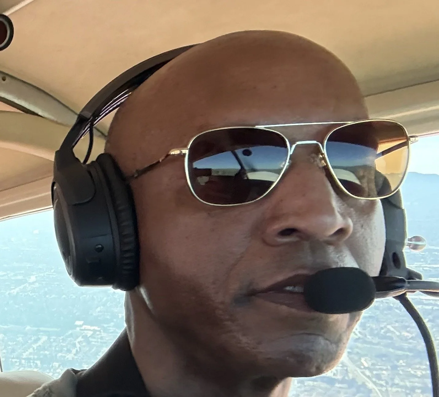 A man wearing sunglasses and a headset with a microphone flying in an aircraft, with a view of the landscape below through the window.