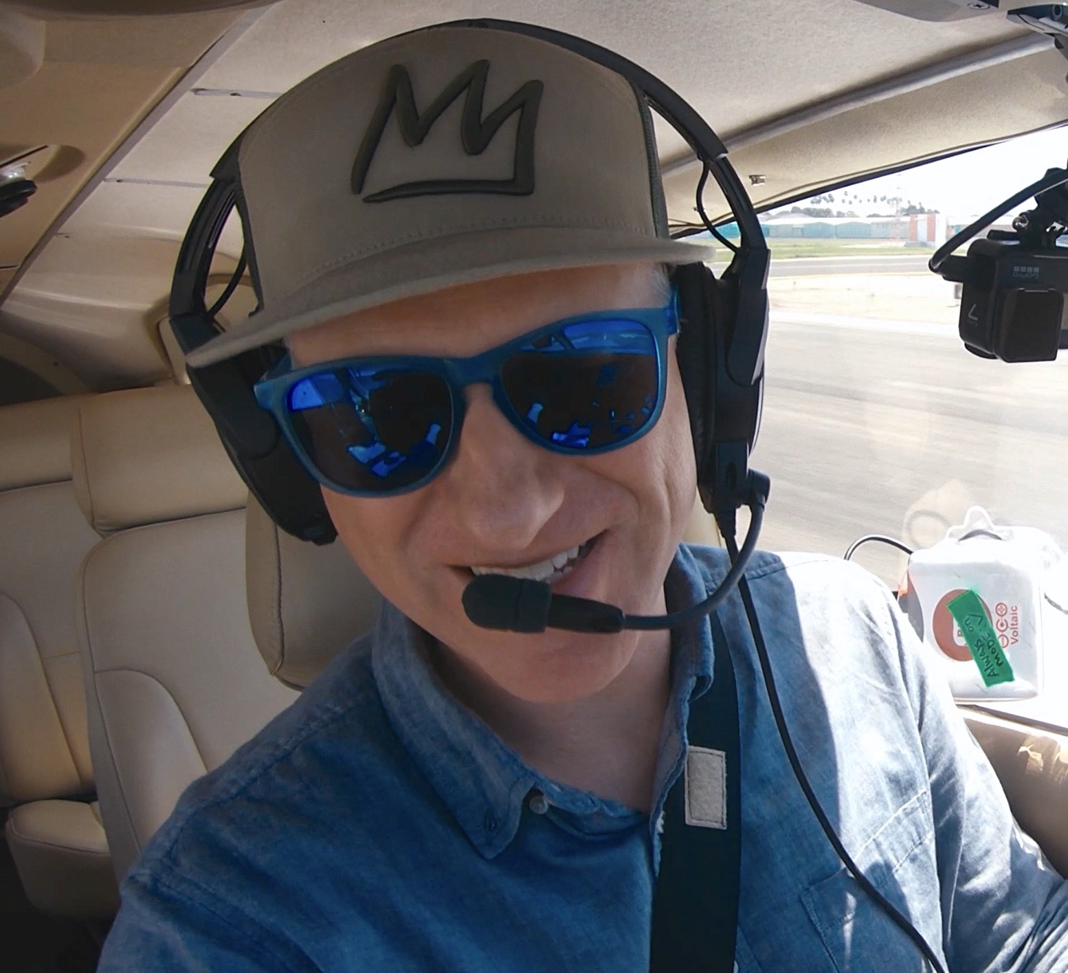 Man wearing a beige cap with a black crown logo, large blue sunglasses, and a headset with a microphone, sitting inside a small aircraft with an aircraft seat and window in the background.
