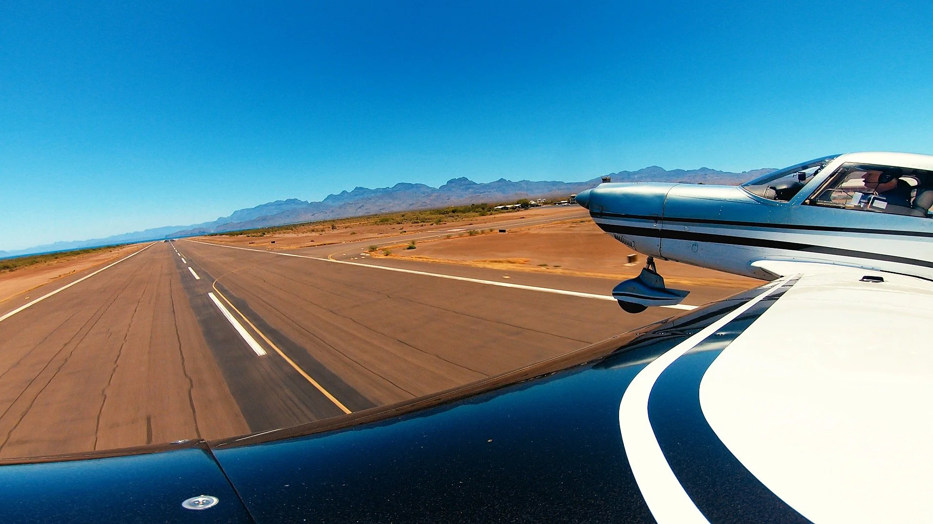 A small airplane flying over a desert runway with mountains in the background under a clear blue sky.