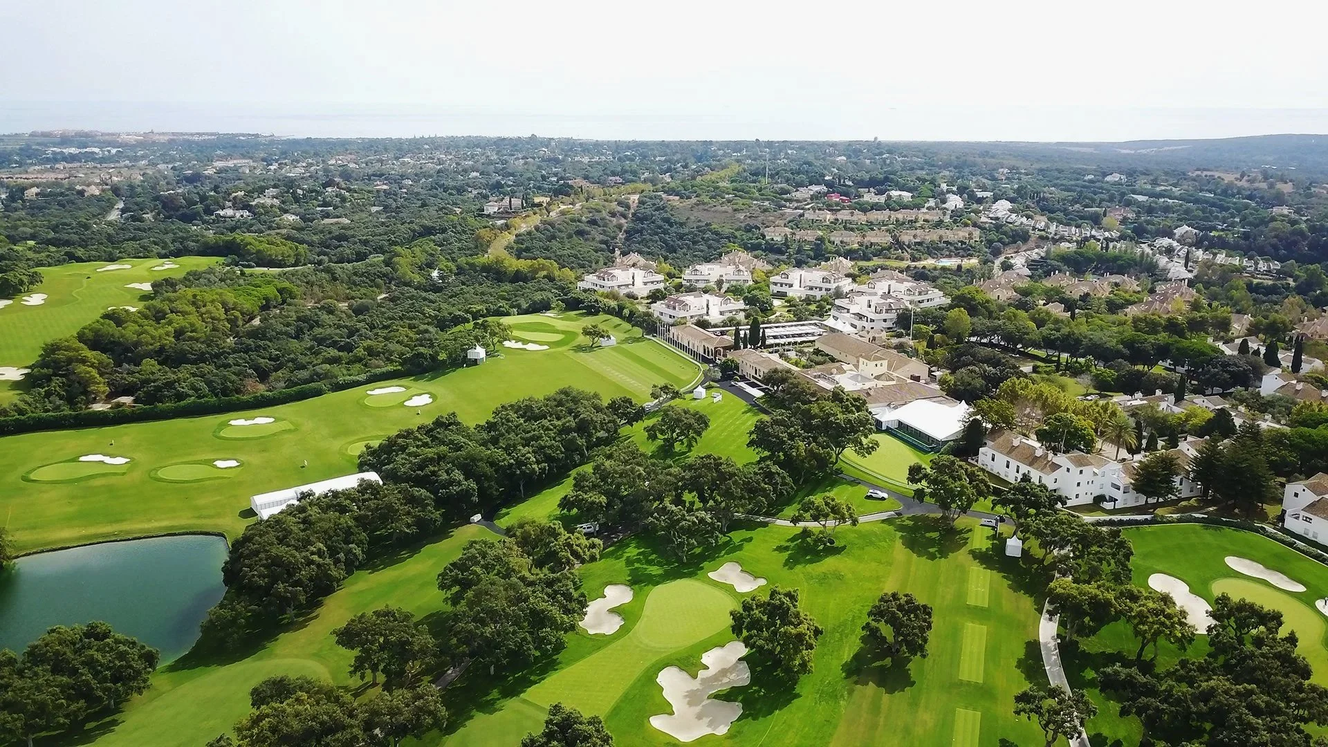Aerial view of a golf course with green fairways, sand traps, a water hazard, and surrounding trees, adjacent to residential houses and neighborhood.