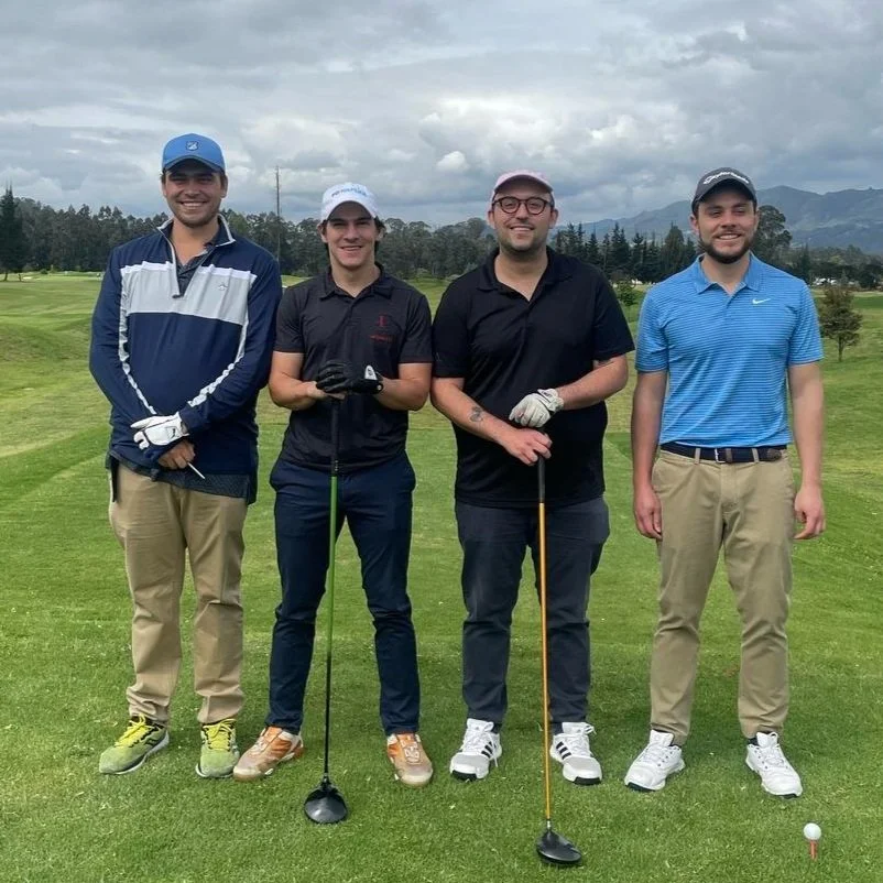 Four men standing on a golf course, smiling, with mountains and cloudy sky in the background.