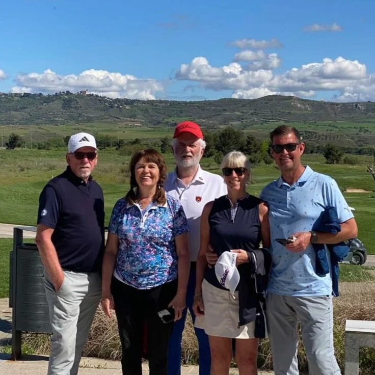 Group of five people standing outdoors on a Spanish golf course, with rolling hills and blue sky with clouds in the background.