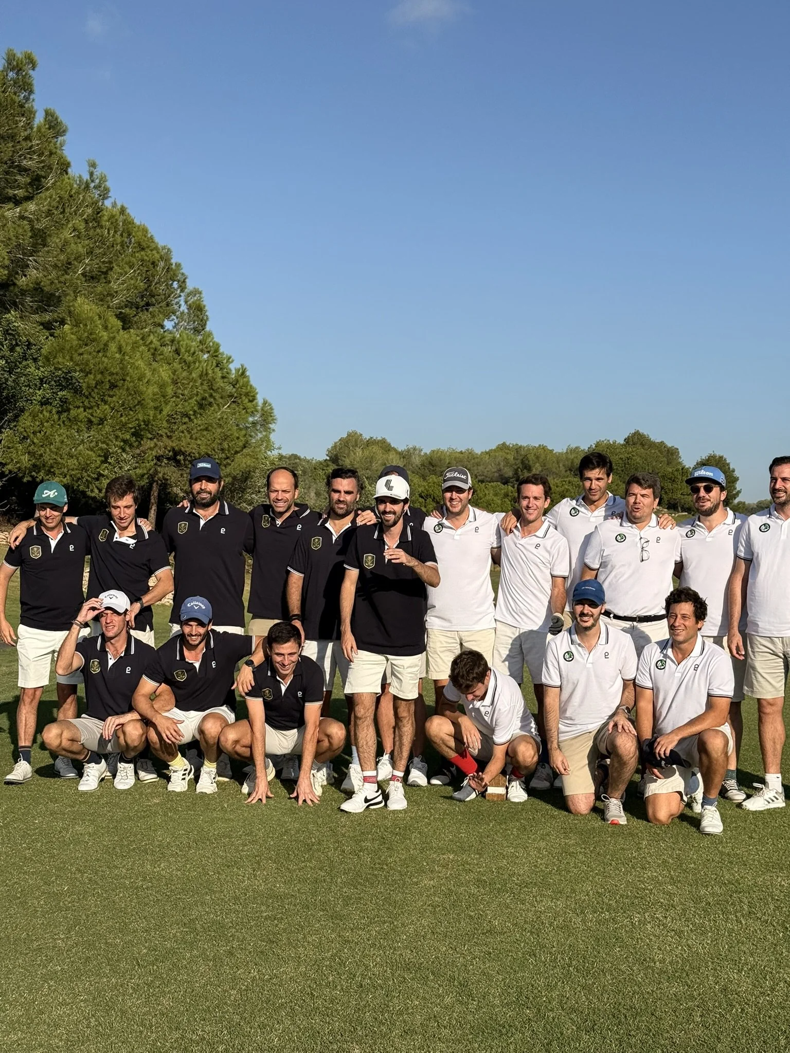 Group of men in golf attire standing and sitting on a golf course under a clear blue sky.
