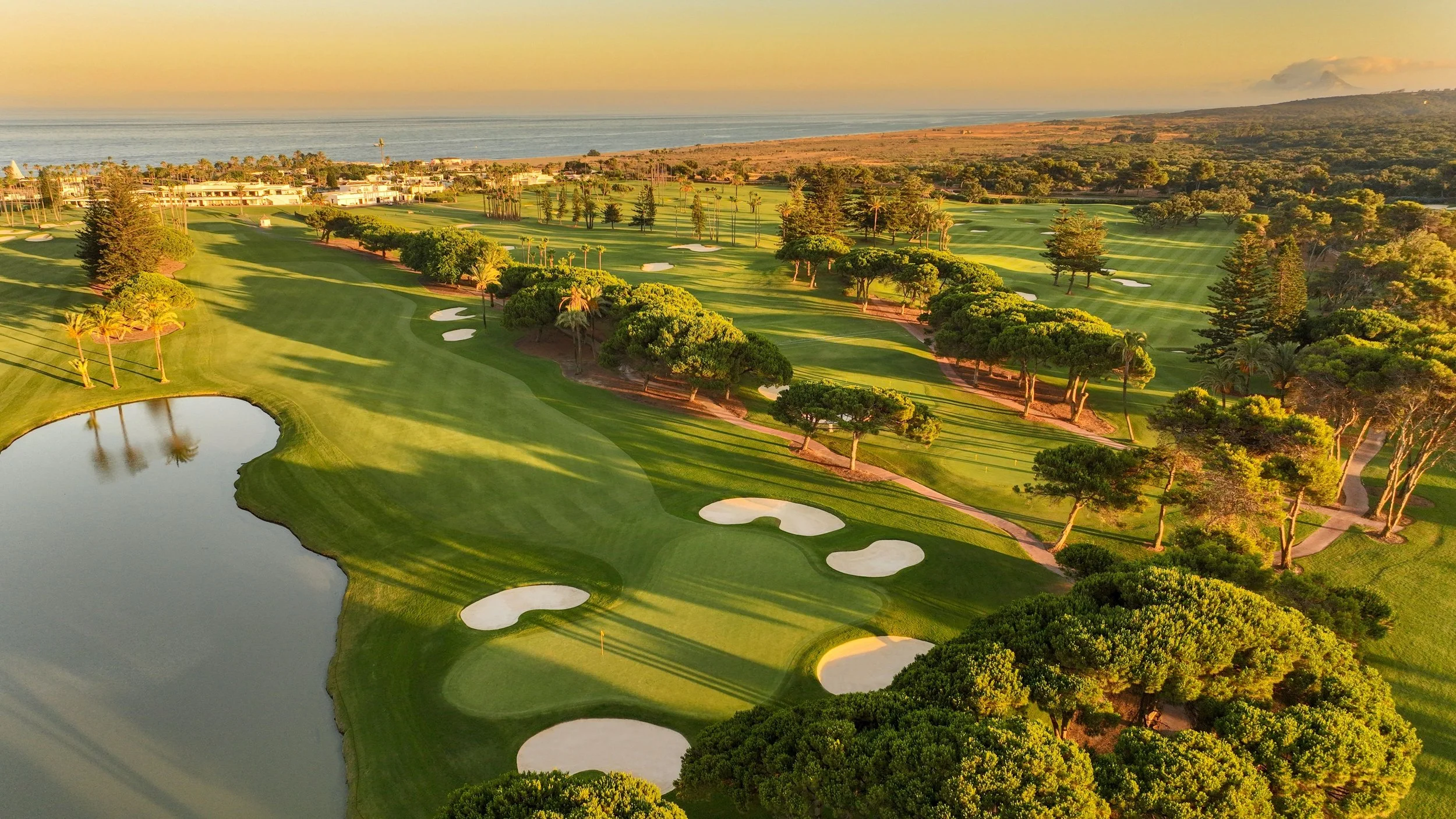 Aerial view of a golf course near the coast with green fairways, sand bunkers, trees, and a large water hazard in the foreground, under a sunset sky.