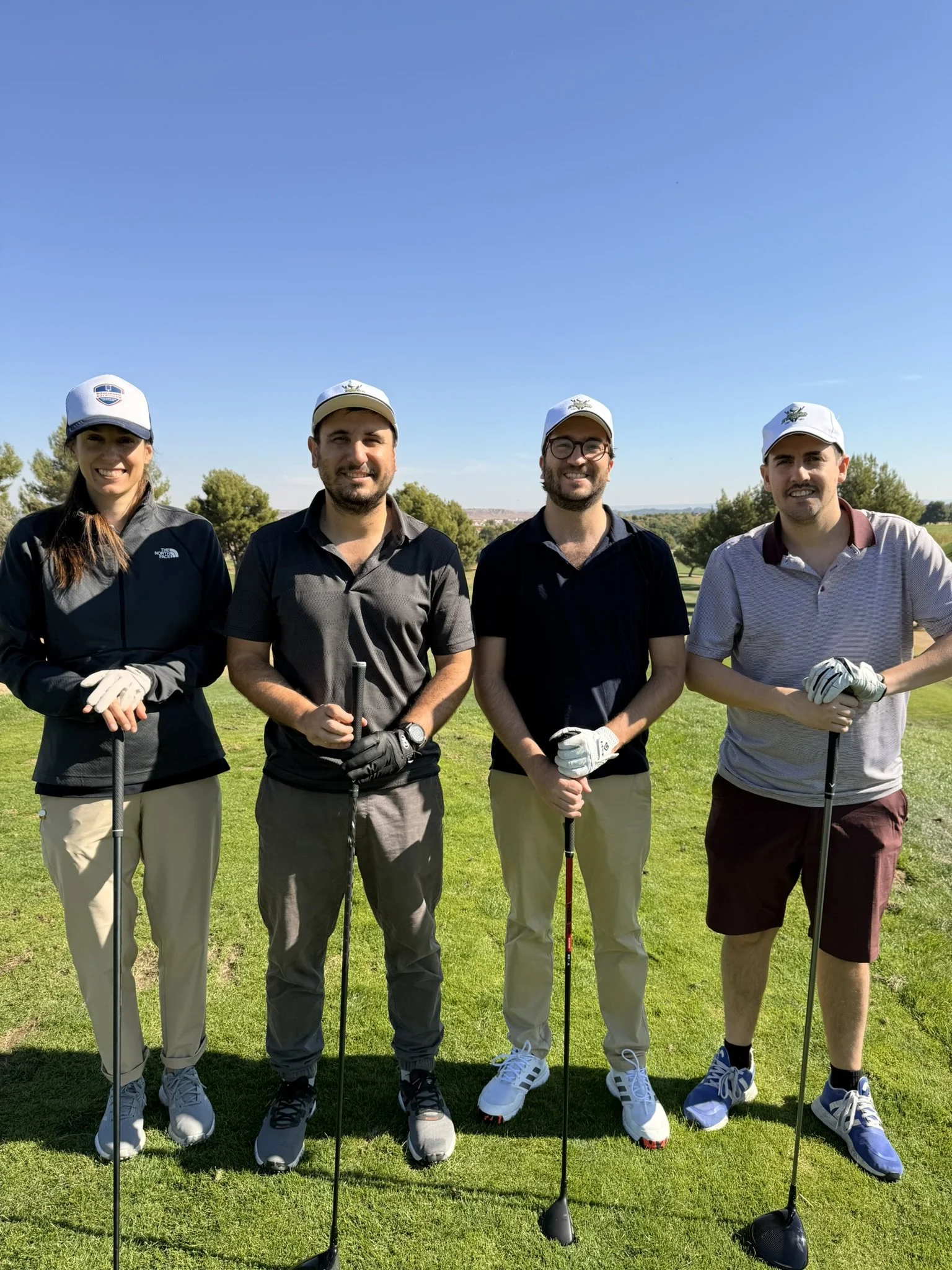 Four people standing on a golf course holding golf clubs, smiling, with trees and a clear blue sky in the background.