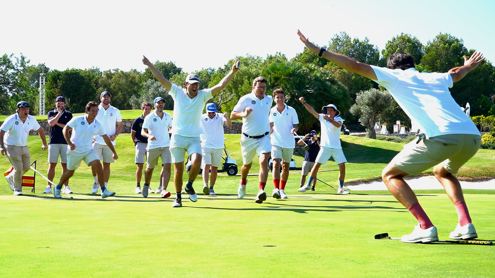 A group of people in white shirts celebrating while playing golf on a sunny day, with one person on the green bending forward with arms outstretched as others cheer.