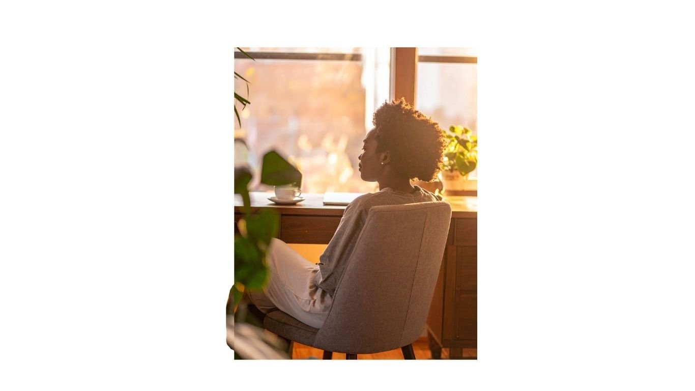 A woman sitting by a window with sunlight illuminating her face and hair, surrounded by houseplants and a wooden table.