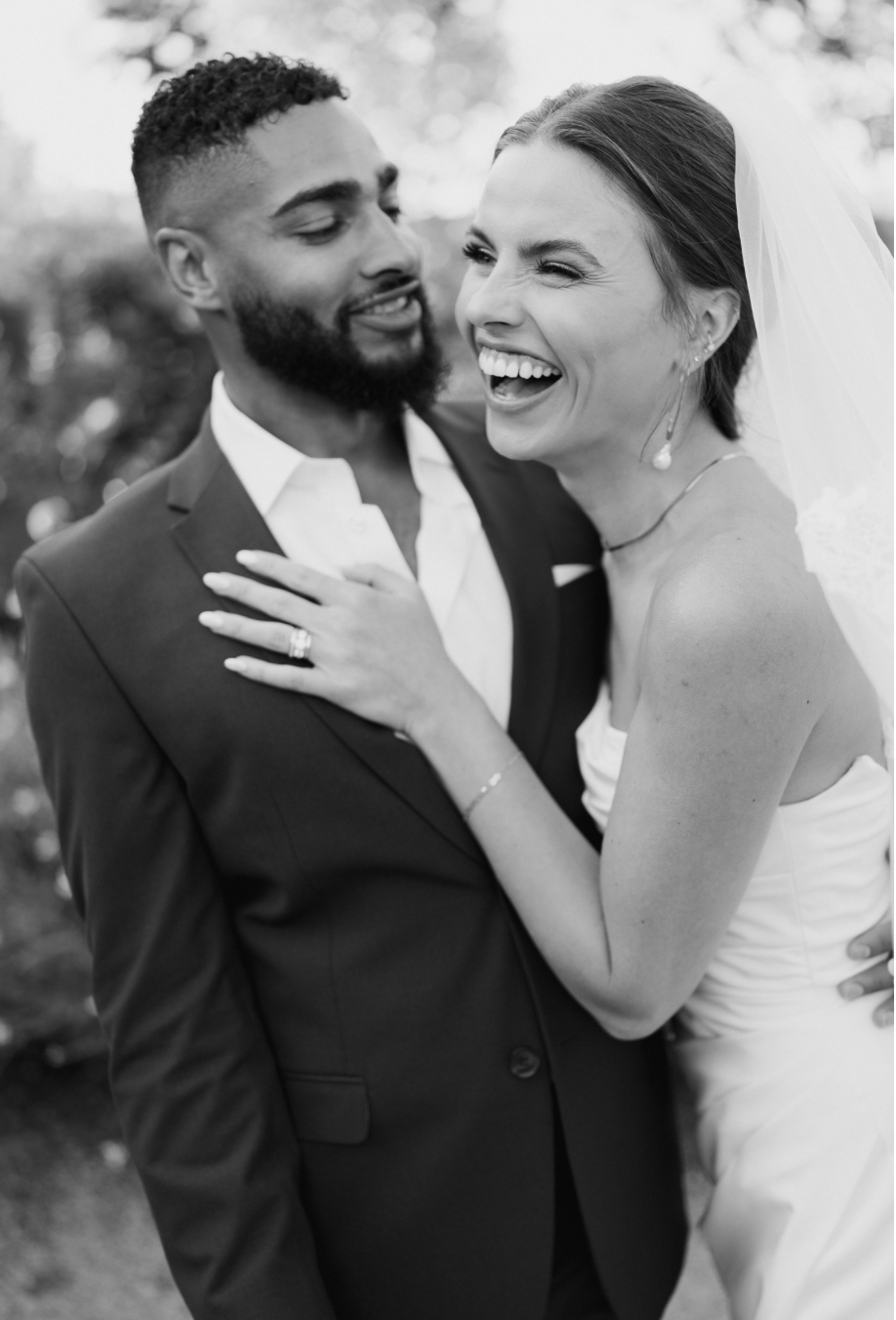 Black and white photo of bride in strapless dress and veil laughing and hugging onto African American groom in suit and open shirt
