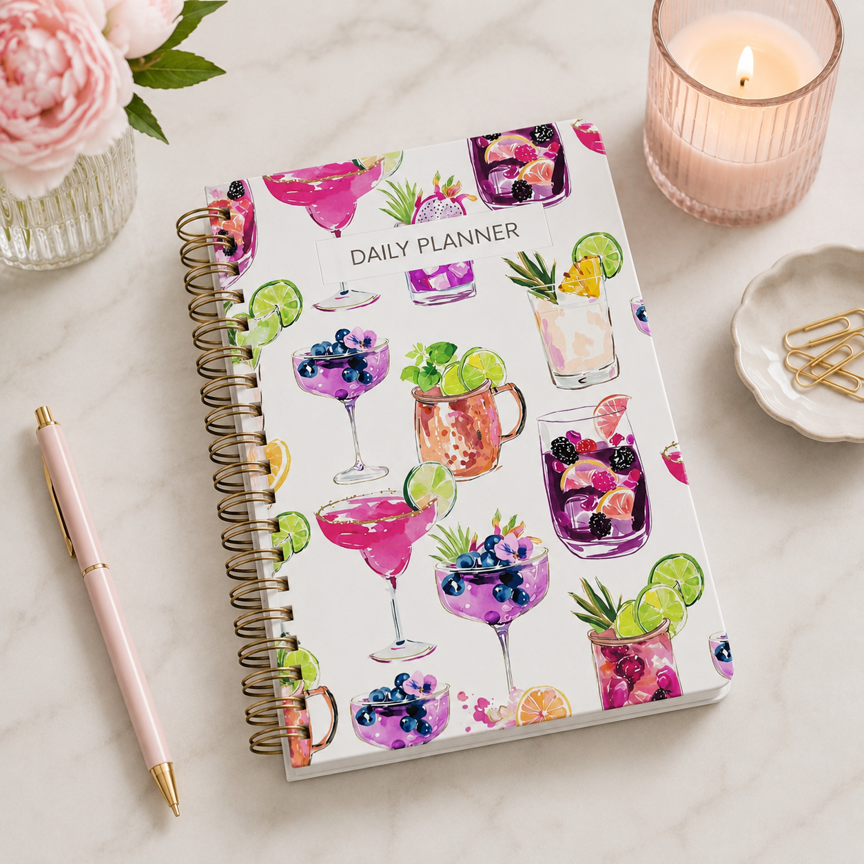 A daily planner with a colorful drink-themed cover design accompanied by a pink pen on a white marble surface. There is a lit pink candle, a small dish with gold paper clips, and a vase of pink flowers nearby.