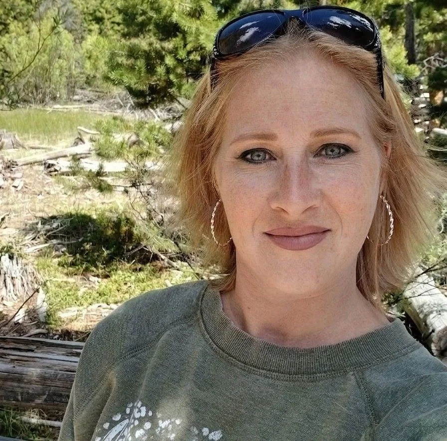 A woman with short red hair, sunglasses on her head, and hoop earrings stands outdoors in a forested area with trees and logs in the background.