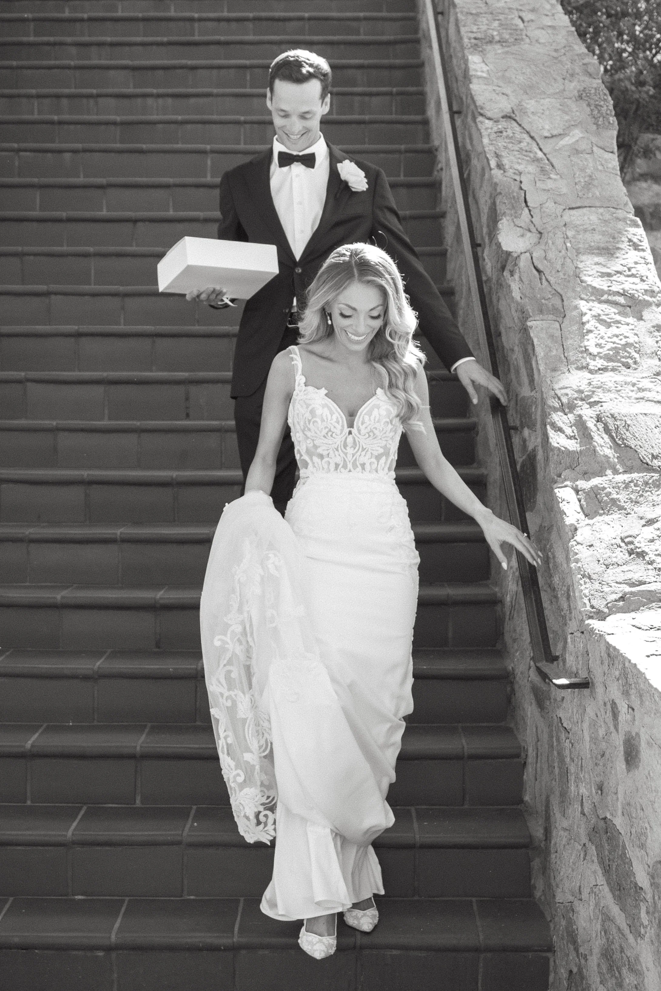 A bride and groom descending outdoor stairs after wedding, with the groom holding a gift box, both smiling.