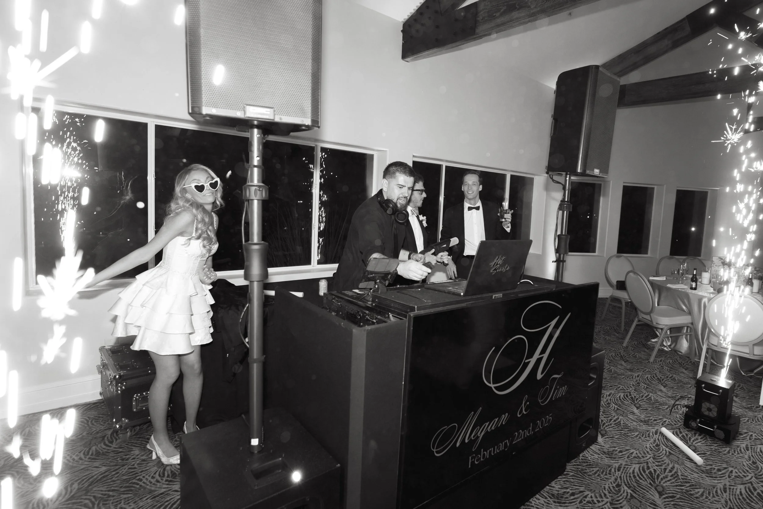 A wedding reception with a DJ, a woman in a ruffled dress and heart-shaped sunglasses, and two men in tuxedos, with table settings and sparklers.