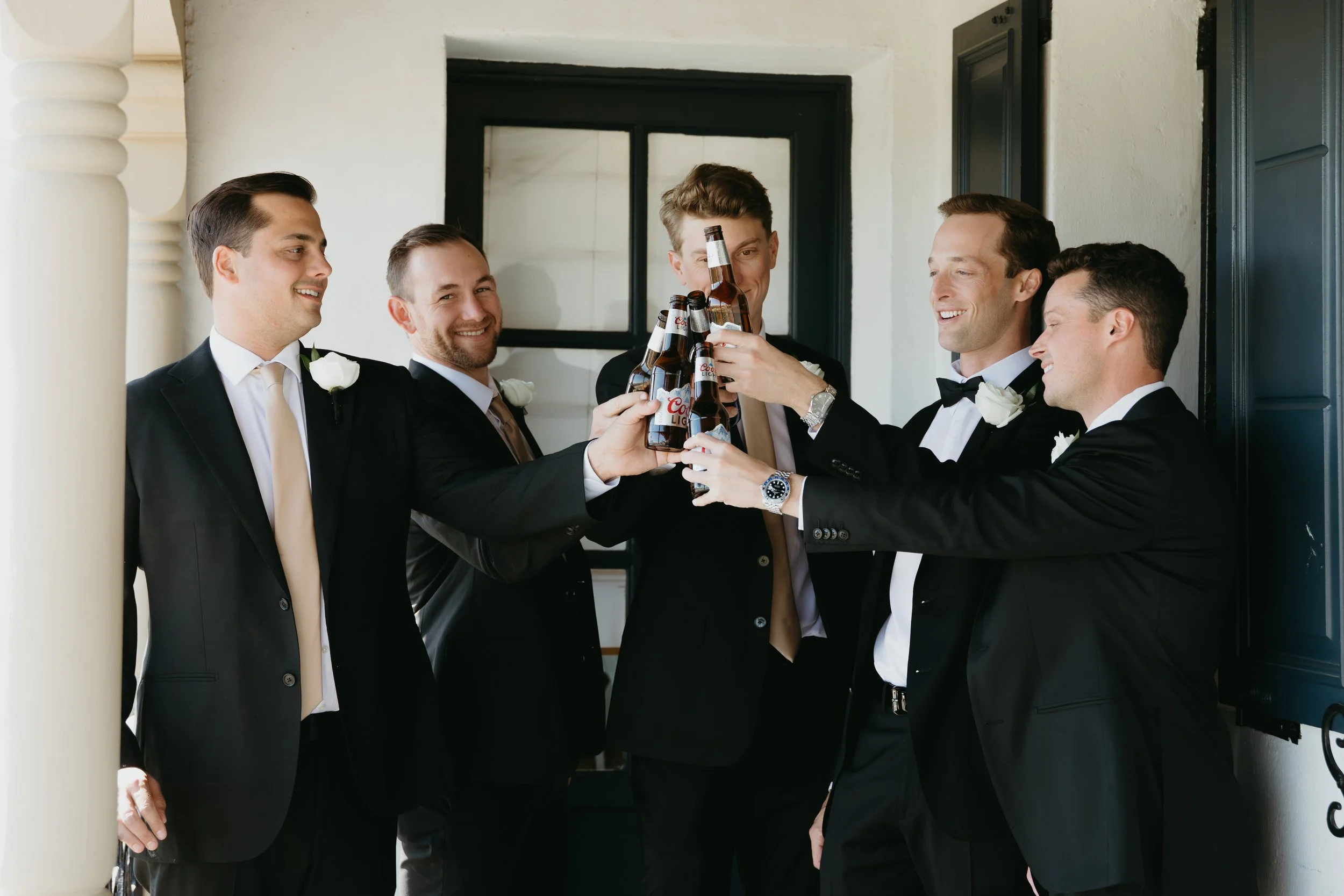 Group of six men in formal tuxedos raising beer bottles in a toast, smiling, in a semi-outdoor setting.