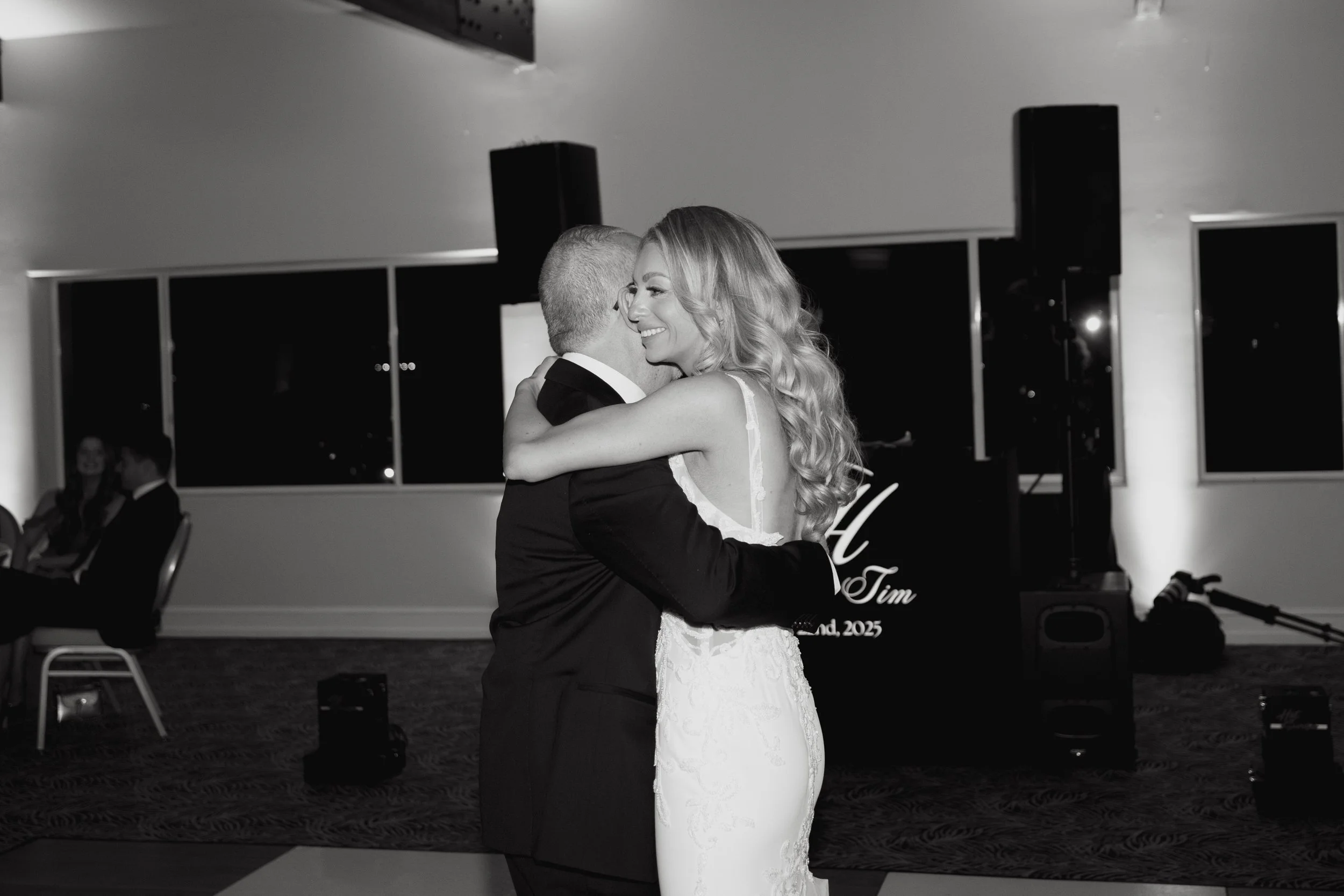 Black and white photo of a bride and groom sharing a dance at their wedding reception, smiling and embracing.