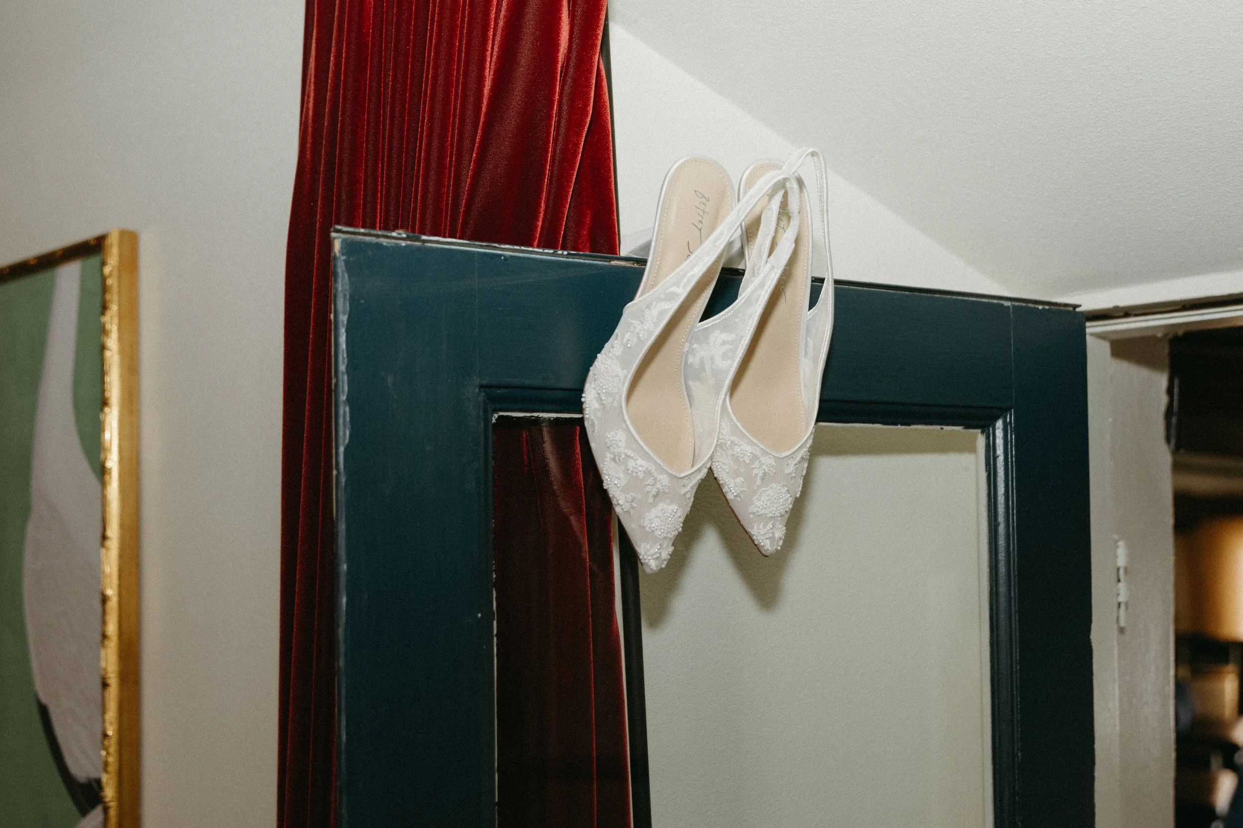 A pair of white, embroidered high-heeled shoes hanging on a black mirror frame with a red velvet curtain in the background.
