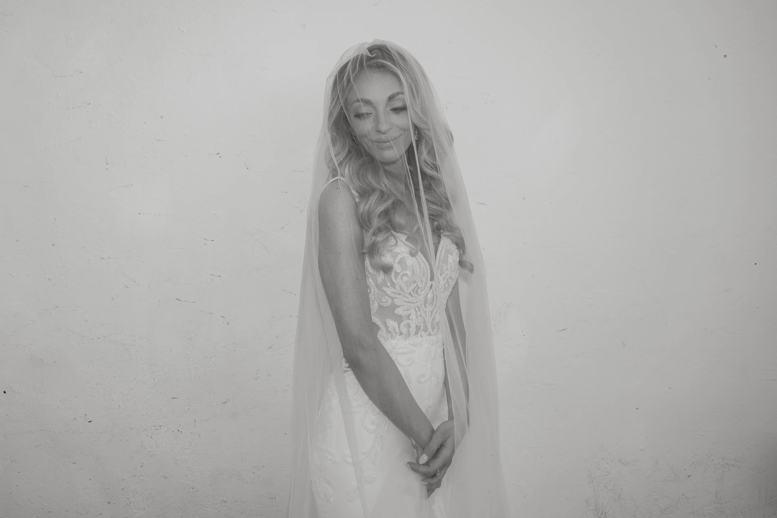 A woman in a wedding dress with a veil, smiling with her eyes closed against a plain white wall.
