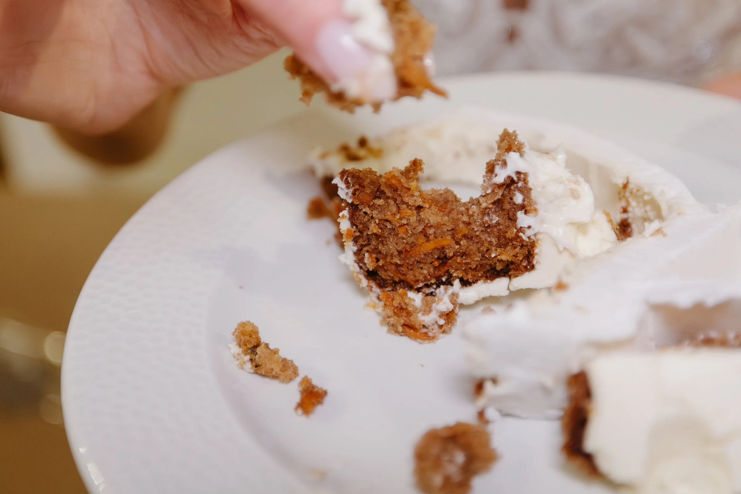 A hand holding a fork with a piece of carrot cake with cream cheese frosting, on a white paper plate.