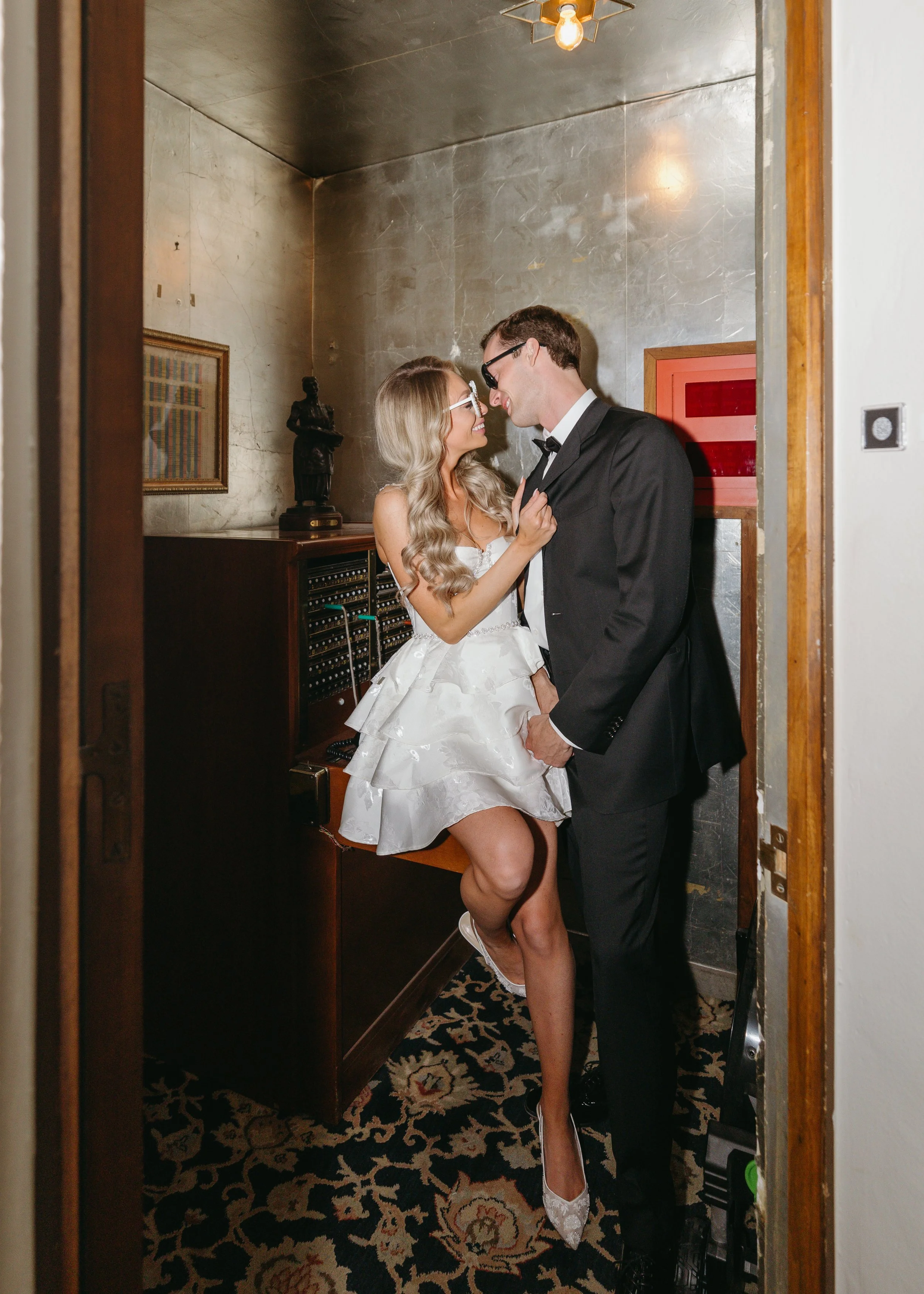 A couple, dressed in wedding attire, sharing an intimate moment in a small room, with the woman sitting on a piano and the man standing in front of her, smiling and touching foreheads.