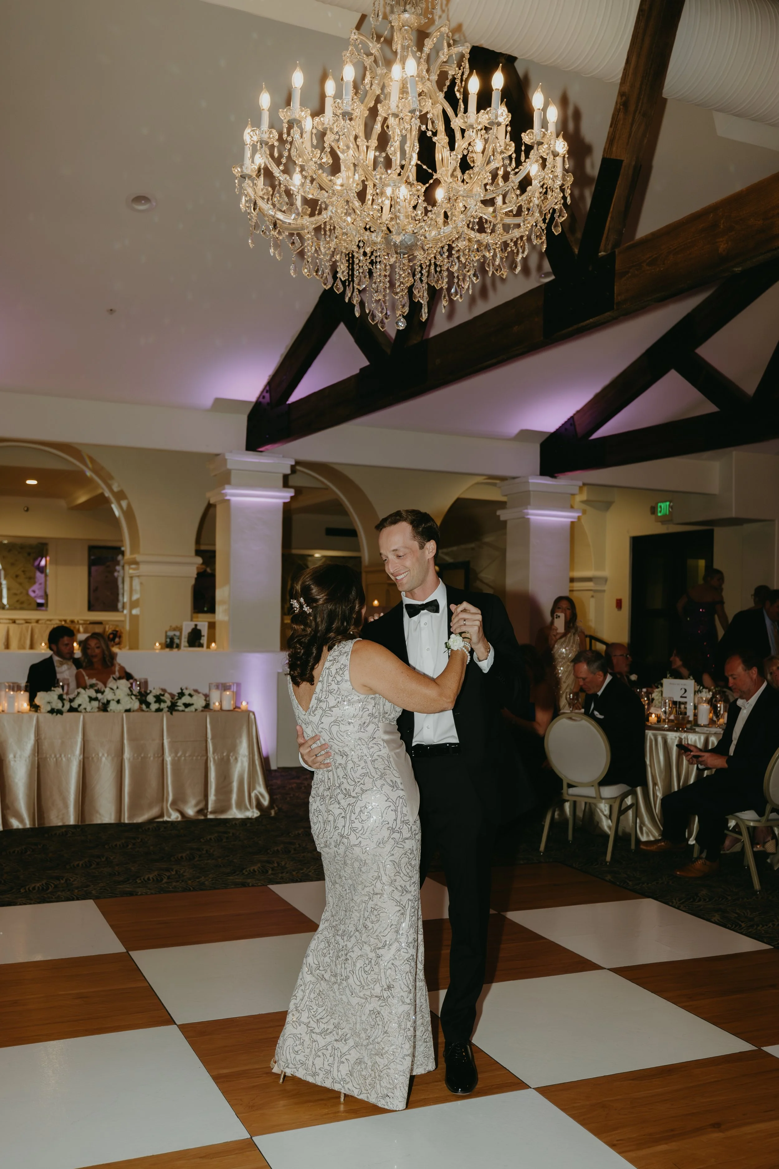 A bride and groom dance together in a wedding reception hall decorated with a chandelier and elegant table settings.