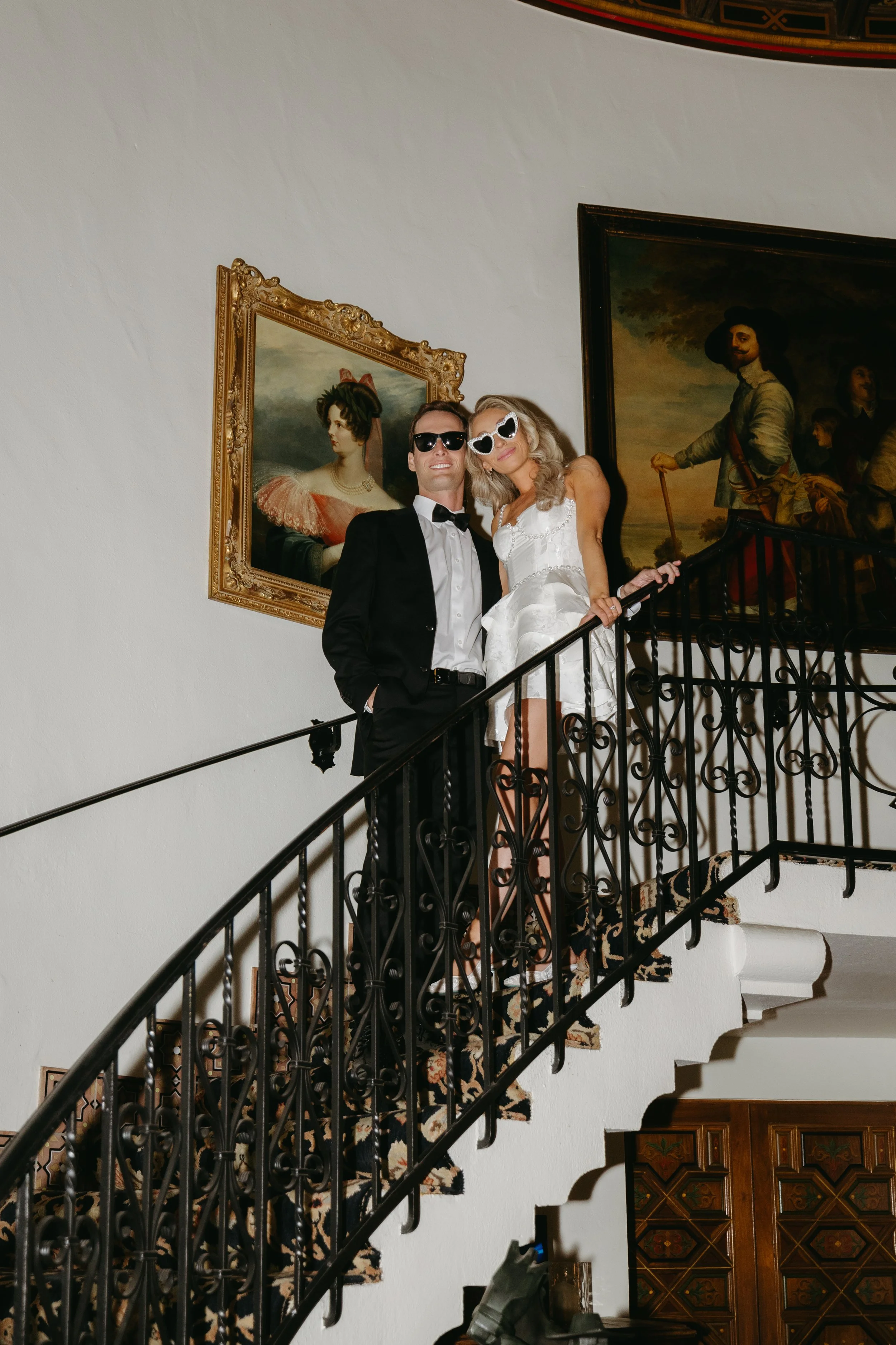 A man in a black tuxedo and a woman in a white dress with sunglasses standing on a staircase inside a building with framed artwork on the wall.