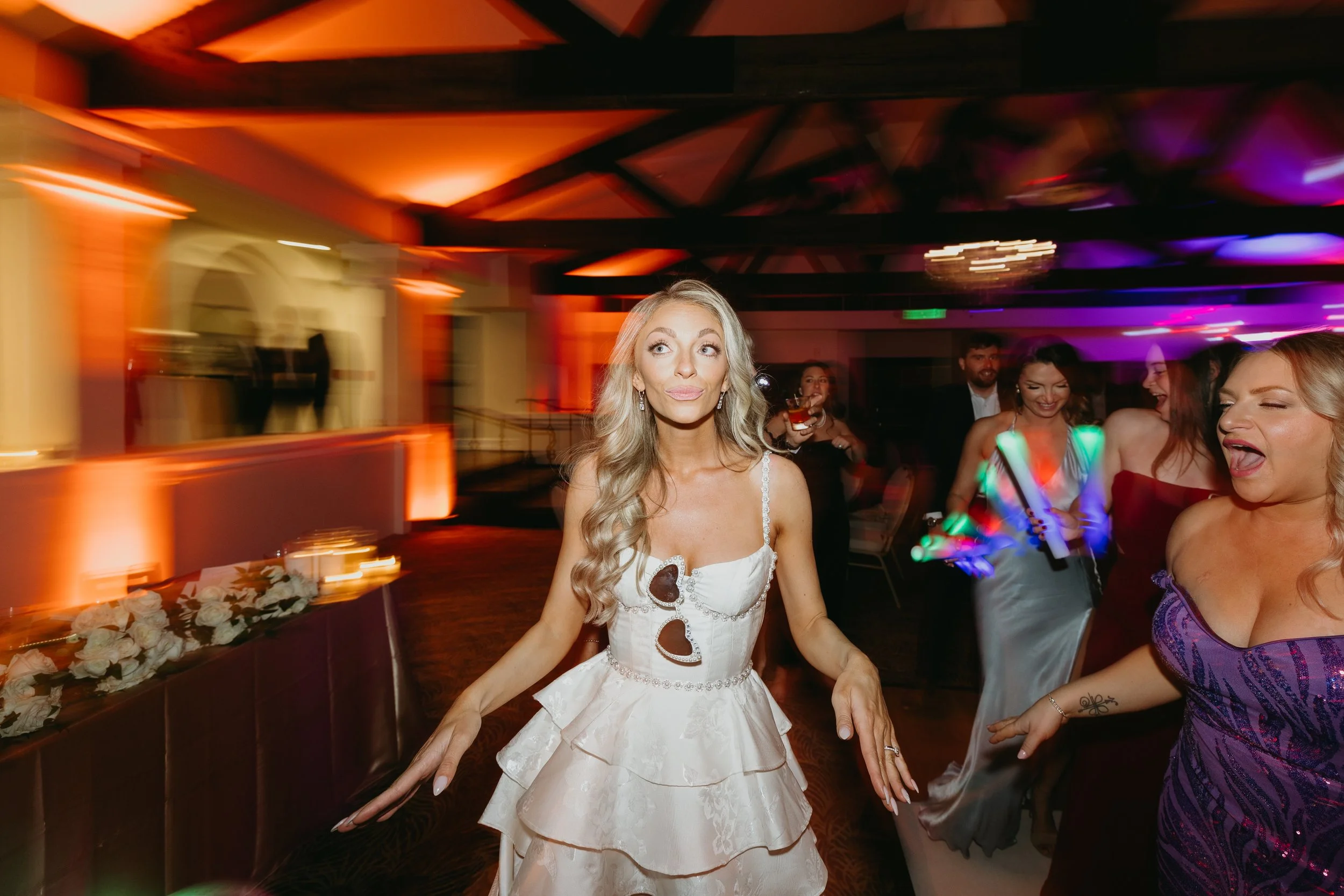 Group of women dancing and celebrating at a party or wedding reception in a dimly lit room with colorful lighting.