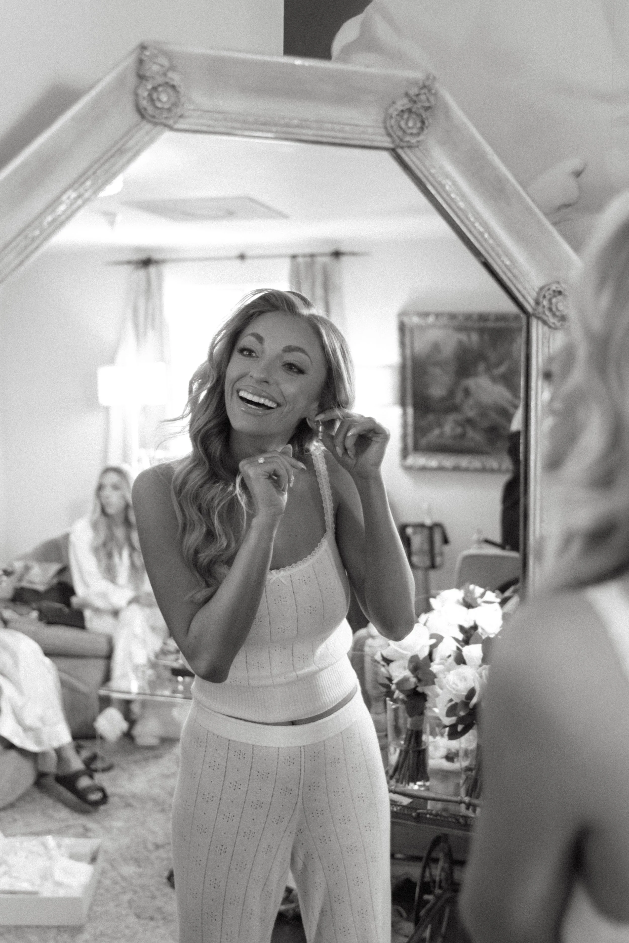 A woman smiling and putting on earrings while looking in a mirror, with another woman sitting on a couch in the background, in a room with floral decor.