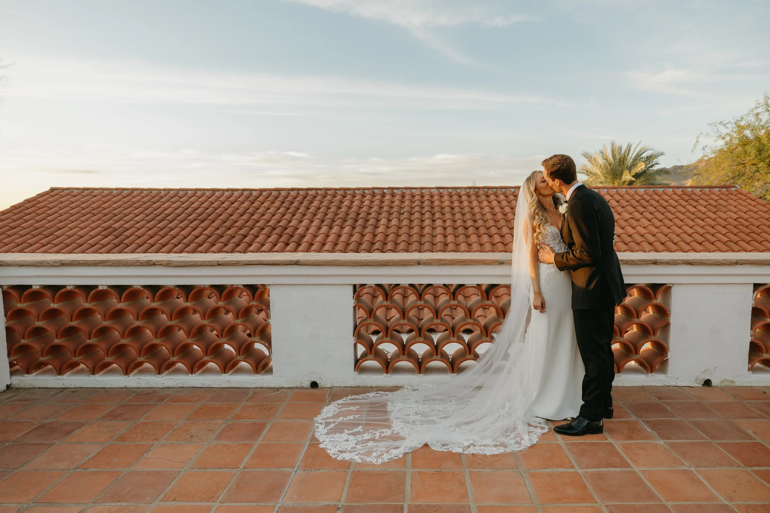 A bride and groom sharing a kiss on a balcony with a terracotta tiled roof and a sunset sky in the background.