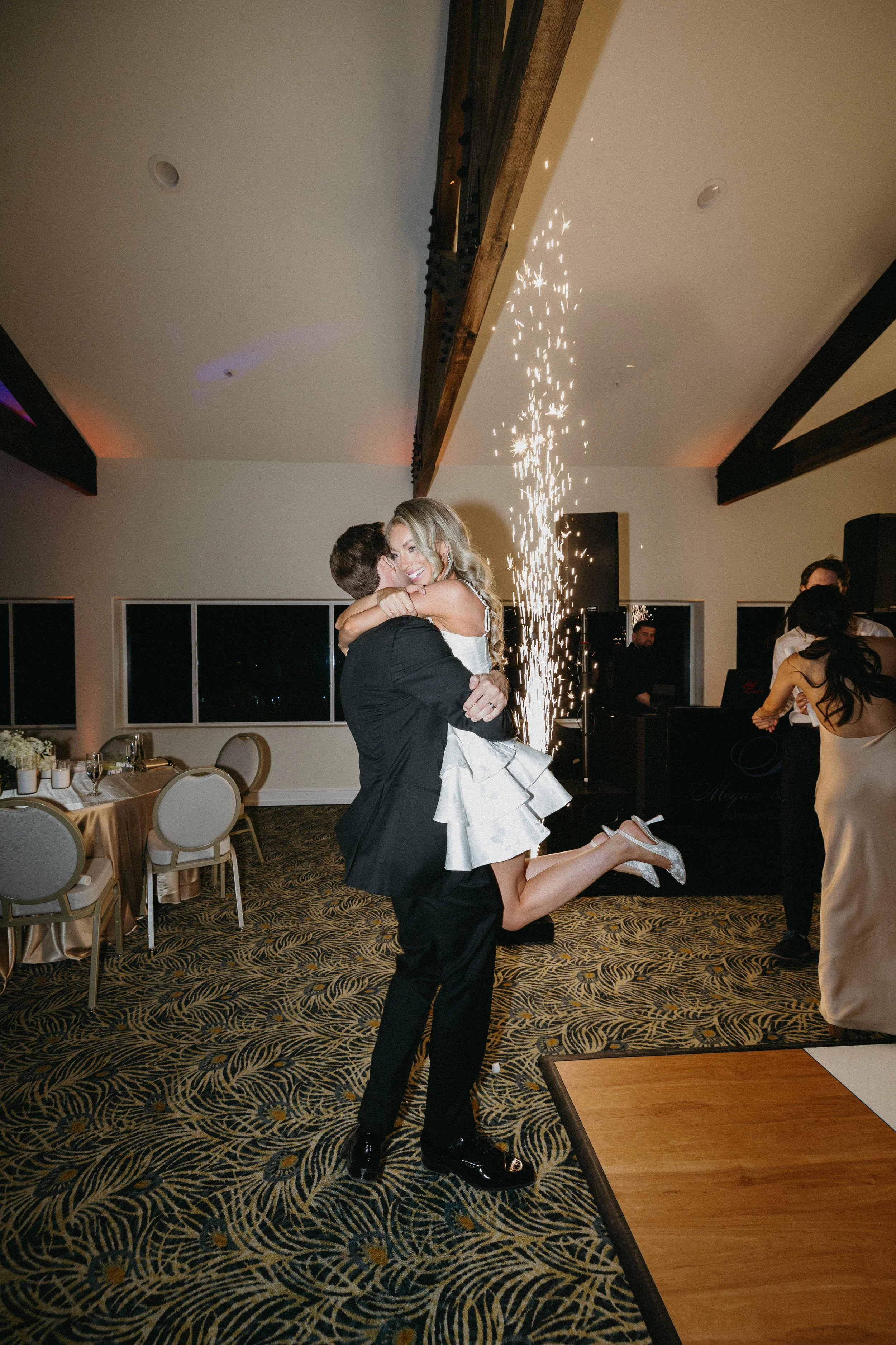 A man in a black suit lifts a woman in a white dress, celebrating with sparklers in the background during a party or wedding reception.