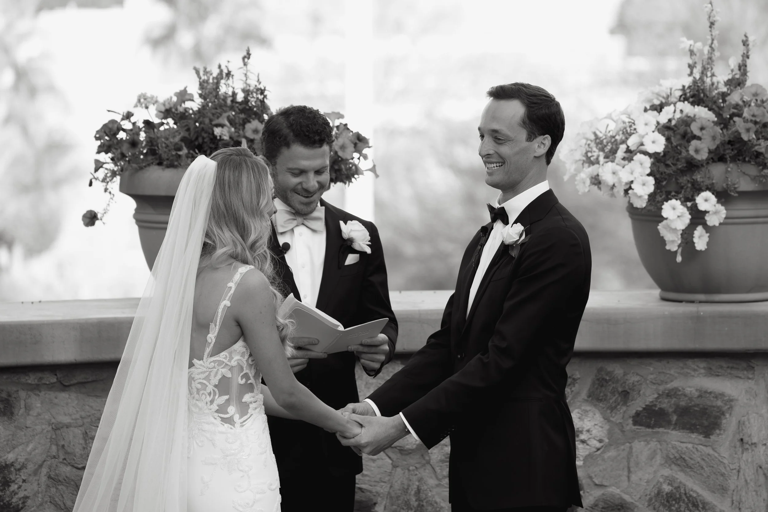 A black-and-white photo of a wedding ceremony where a bride and groom are holding hands, smiling, and facing each other, with an officiant reading from a book. Large flower arrangements are visible in the background.