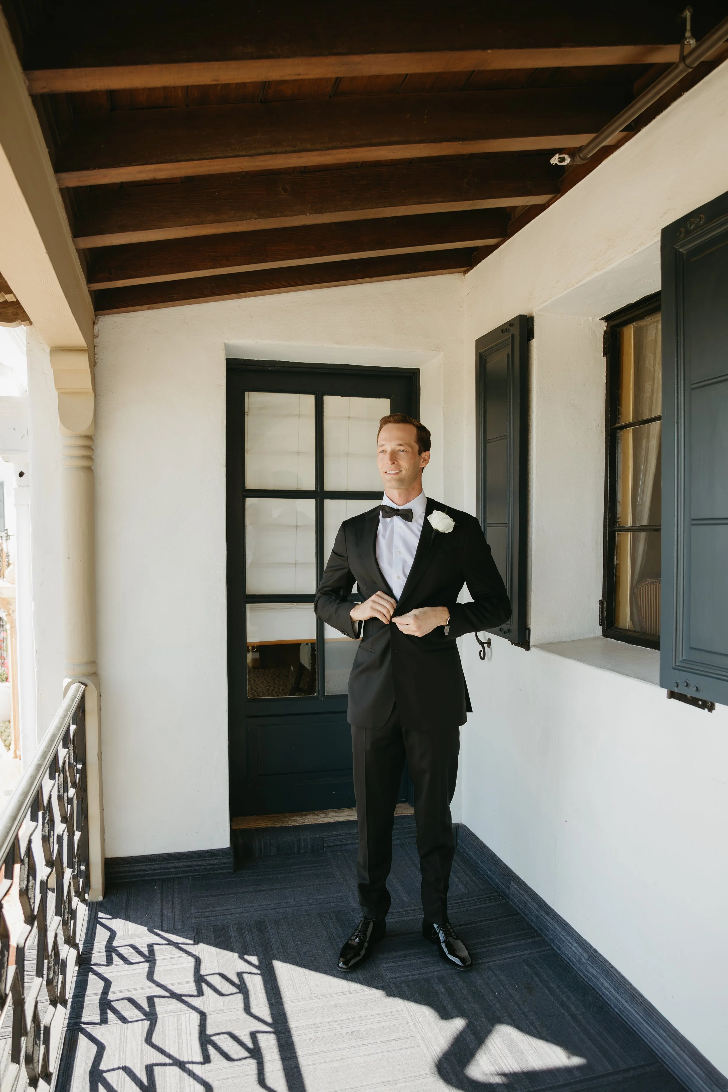 A groom dressed in a black tuxedo with a bow tie and white flower boutonniere standing on a balcony, adjusting his jacket, smiling, with black window shutters and a wooden ceiling in the background.