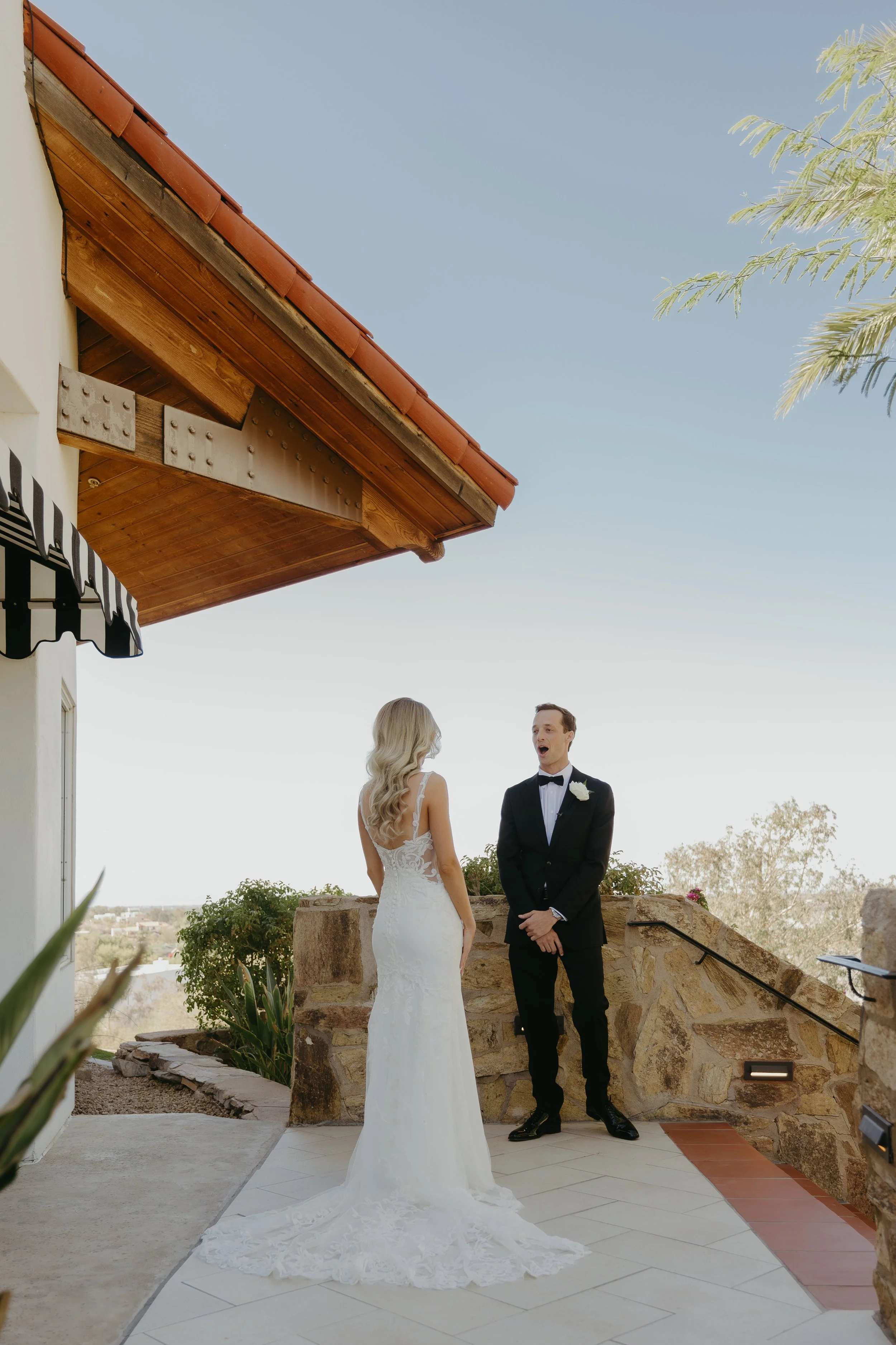 A bride and groom standing outside on a stone patio, facing each other, during their wedding. The bride is in a white lace wedding dress with a train, and the groom is in a black tuxedo with a bow tie and white boutonniere. The background shows a cle