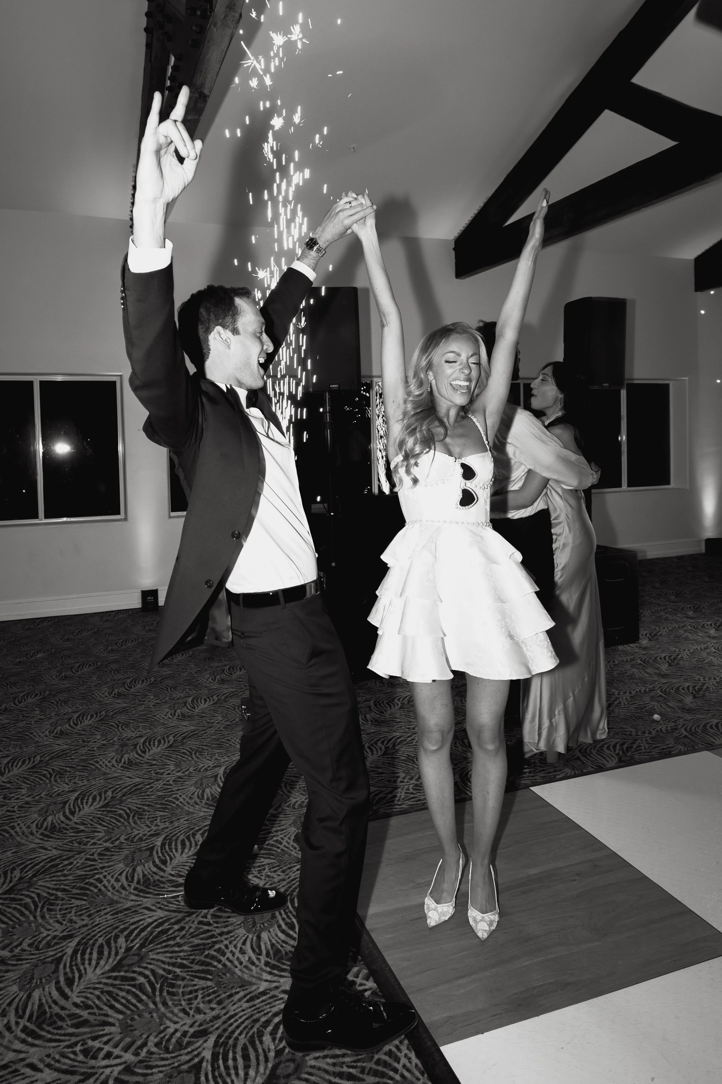 A man and woman dancing happily at a celebration or party, with the man holding sparklers and the woman with her arms raised.