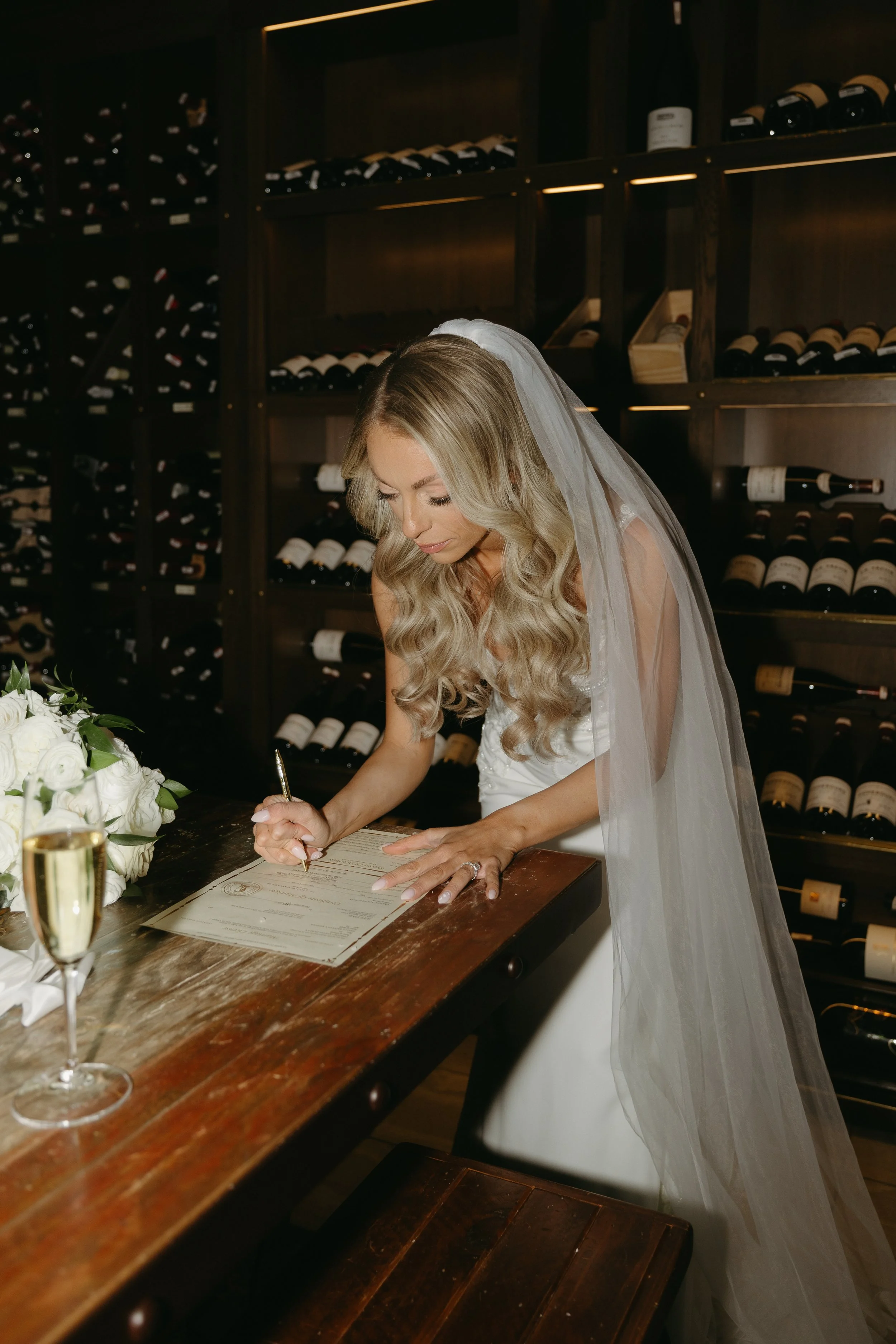 Bride signing a marriage certificate in a wine cellar with wine bottles on wooden shelves and a glass of champagne on the table.