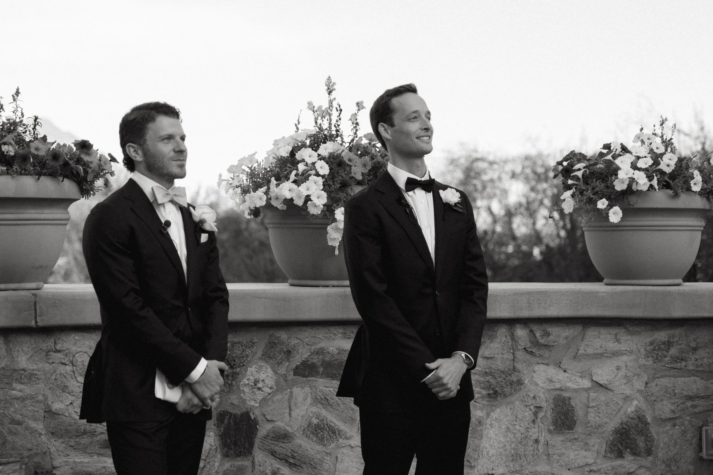 Two men in tuxedos at a wedding ceremony, standing outdoors in front of a stone wall with large flower pots filled with flowers behind them, black and white photo.