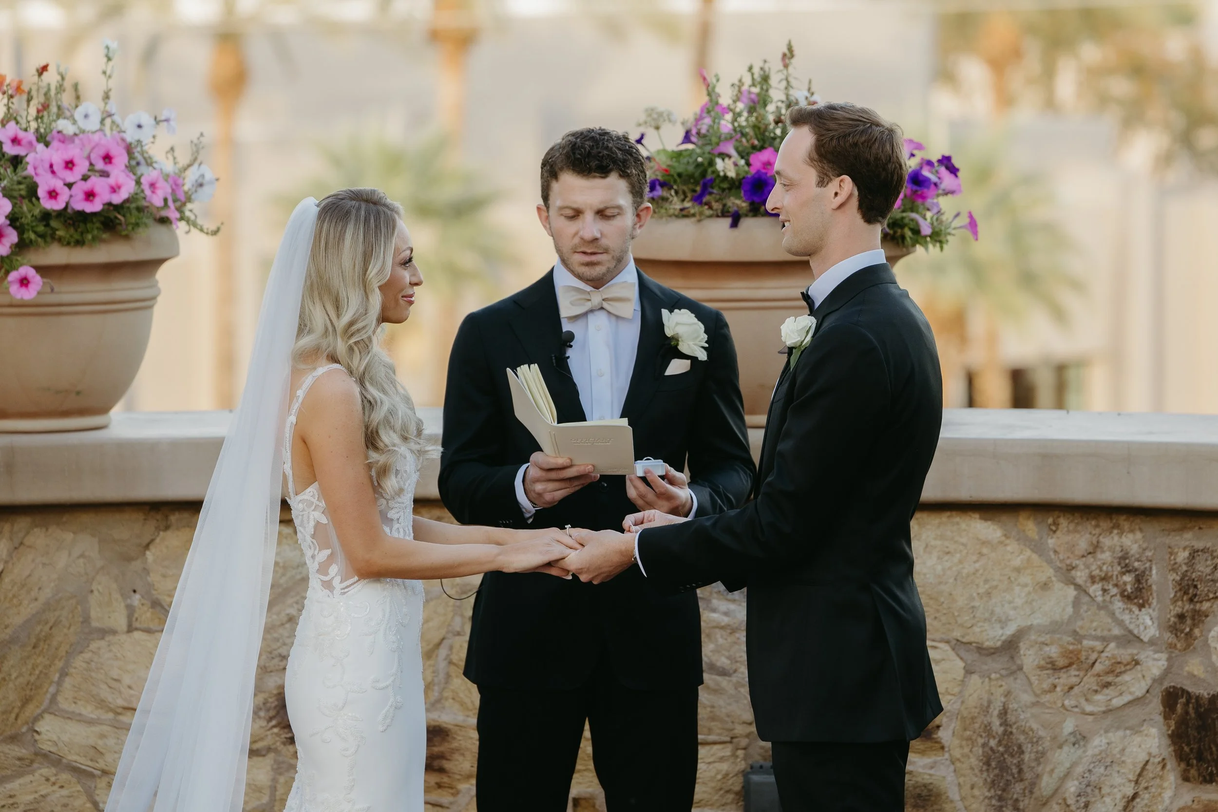 A bride and groom holding hands during their outdoor wedding ceremony, with an officiant reading from a book. The bride wears a white lace wedding dress and veil, and the groom wears a black tuxedo. Large flower pots with pink and purple flowers are
