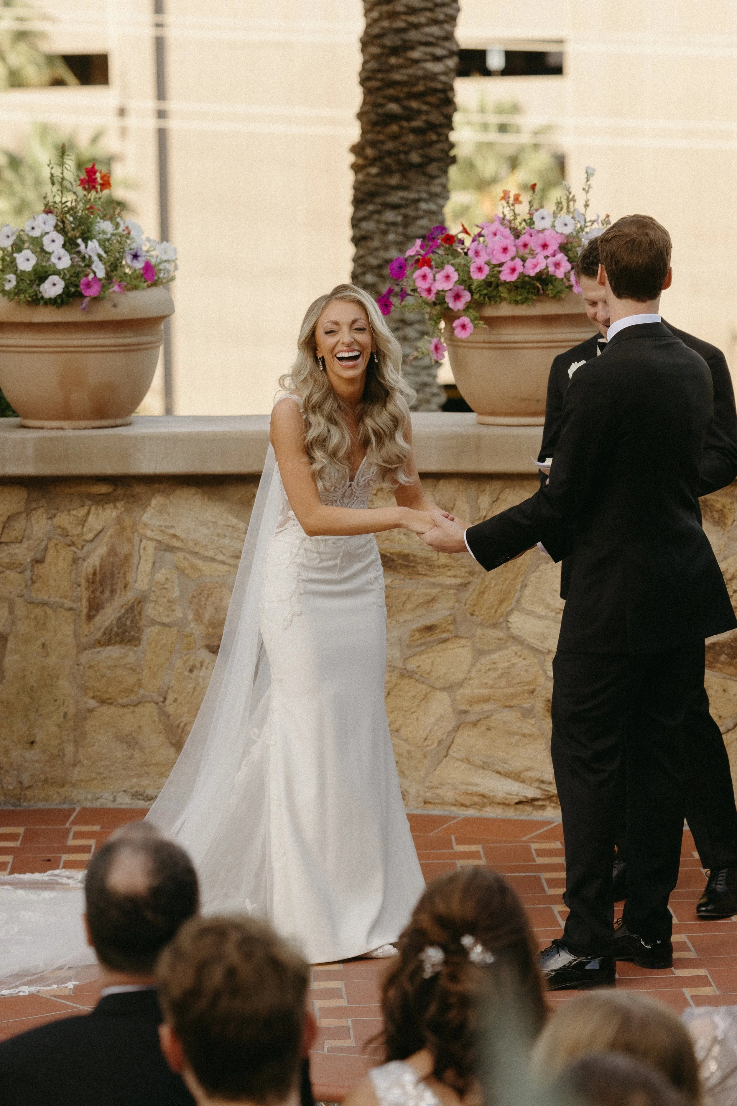 A bride in a white wedding dress and a groom in a black tuxedo holding hands and smiling, exchanging vows during a wedding ceremony outdoors, with guests seated in front and pink and purple flowers in large pots behind them.