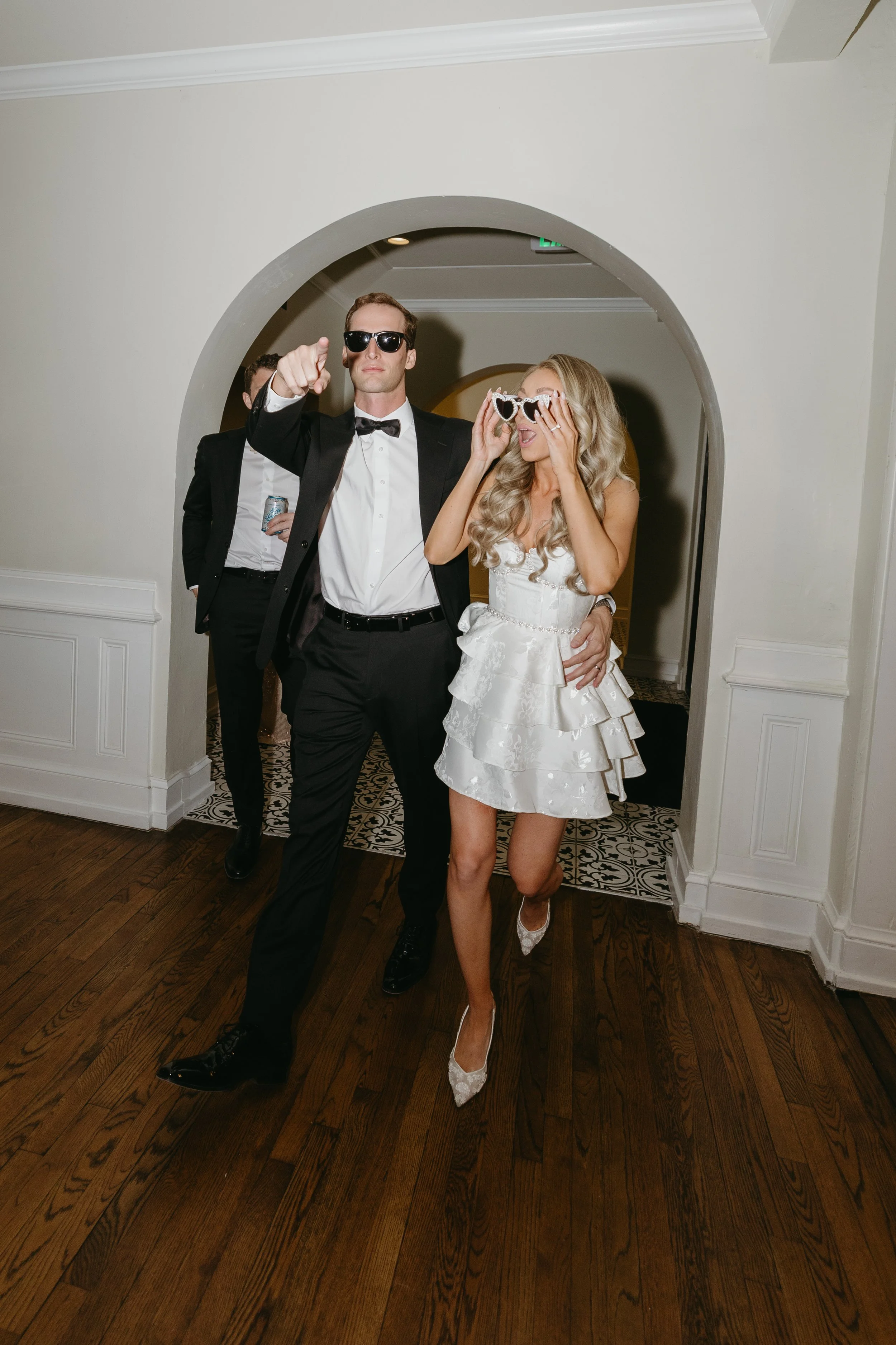 People dressed in formal attire at a party, with one woman wearing heart-shaped sunglasses, and a man pointing at the camera.