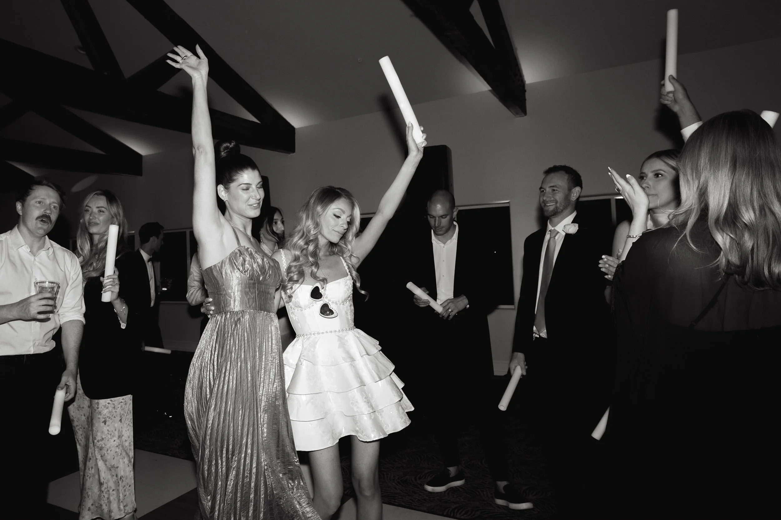 People dancing and celebrating at a party, some holding rolled-up papers or scrolls, in a dimly lit room.