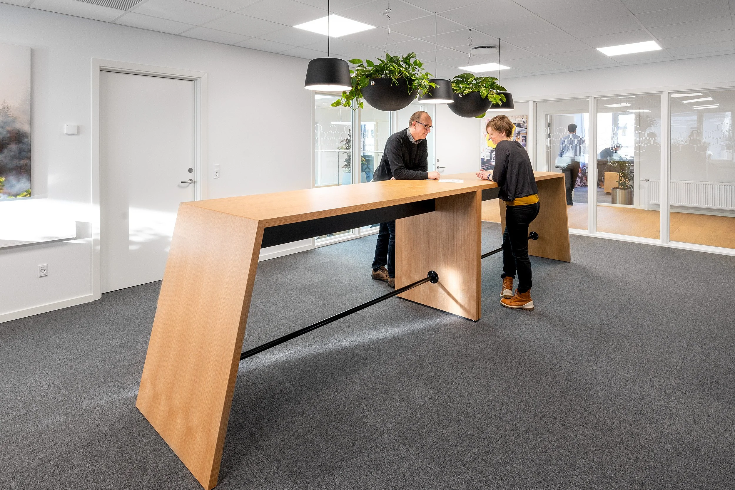 To personer står ved en trædisk, der fungerer som en frontdesk i et moderne kontormiljø, med grønne planter hængende i sorte potter over dem.