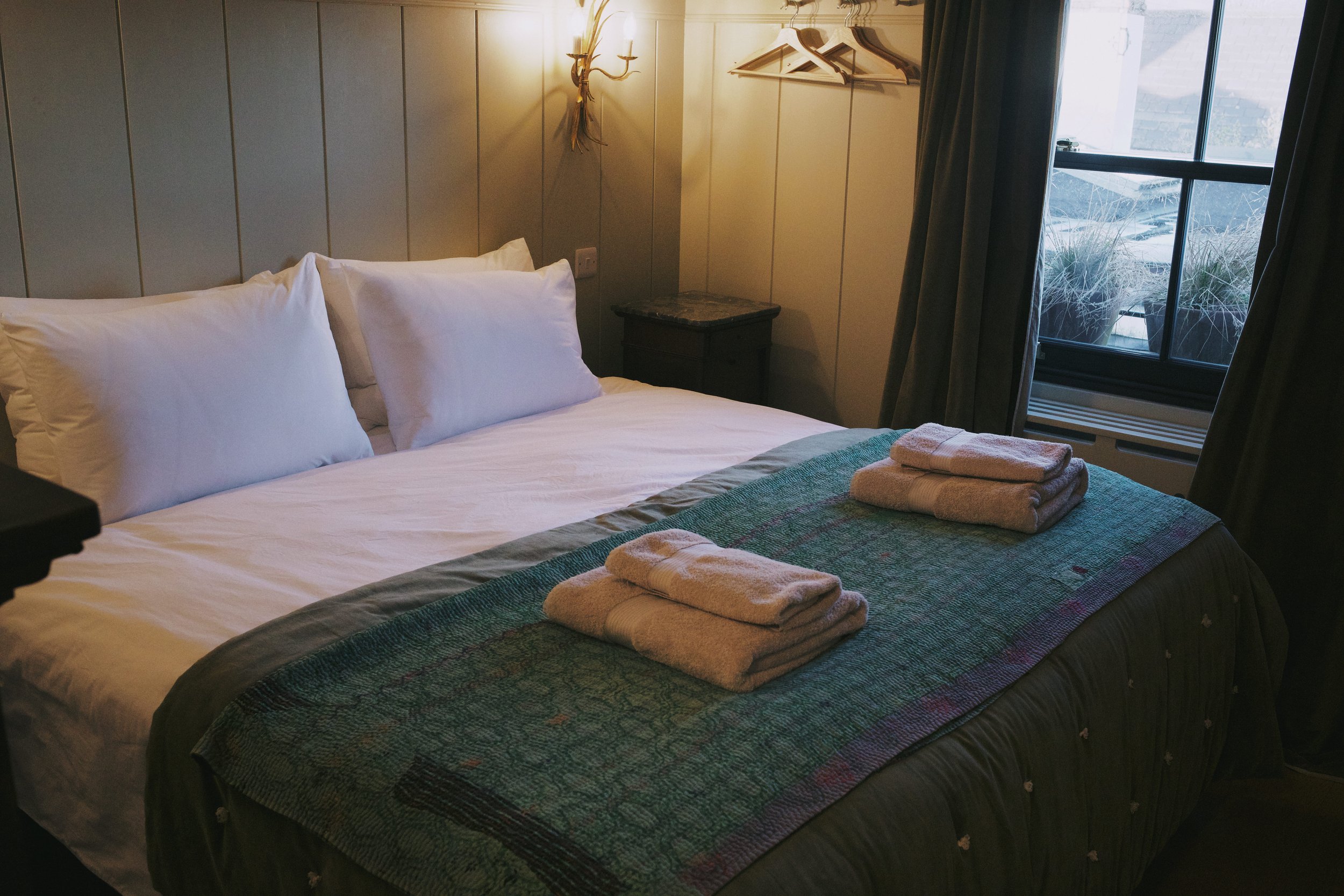 A neatly made bed with white pillows, a colorful quilt, and folded towels on top. A window with dark curtains shows outdoor potted plants, and a wall light fixture is on the headboard.