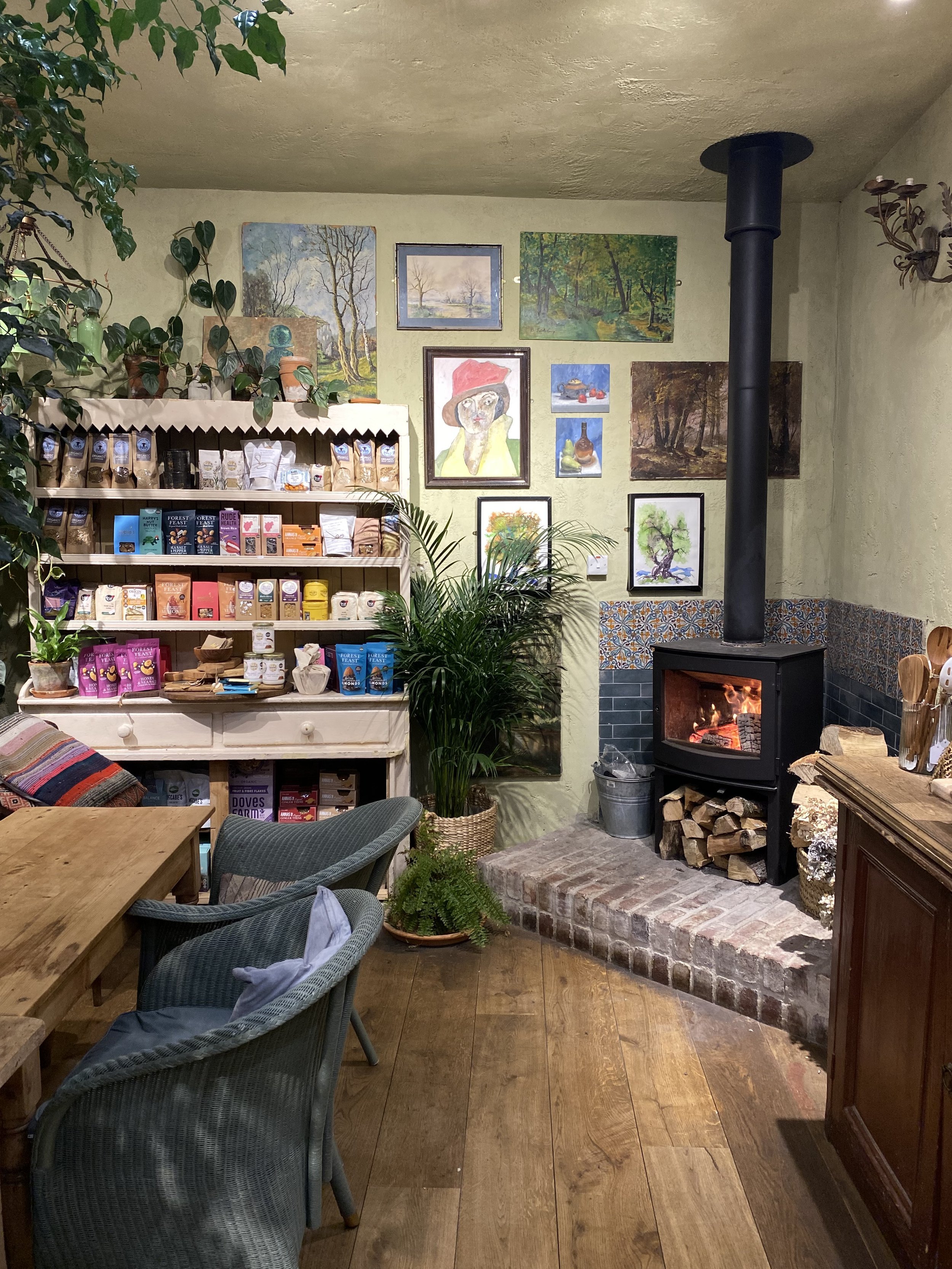 Cozy room with a wood stove, a wooden table with chairs, a bookshelf filled with packaged foods, and wall art above it. There are plants and warm lighting.