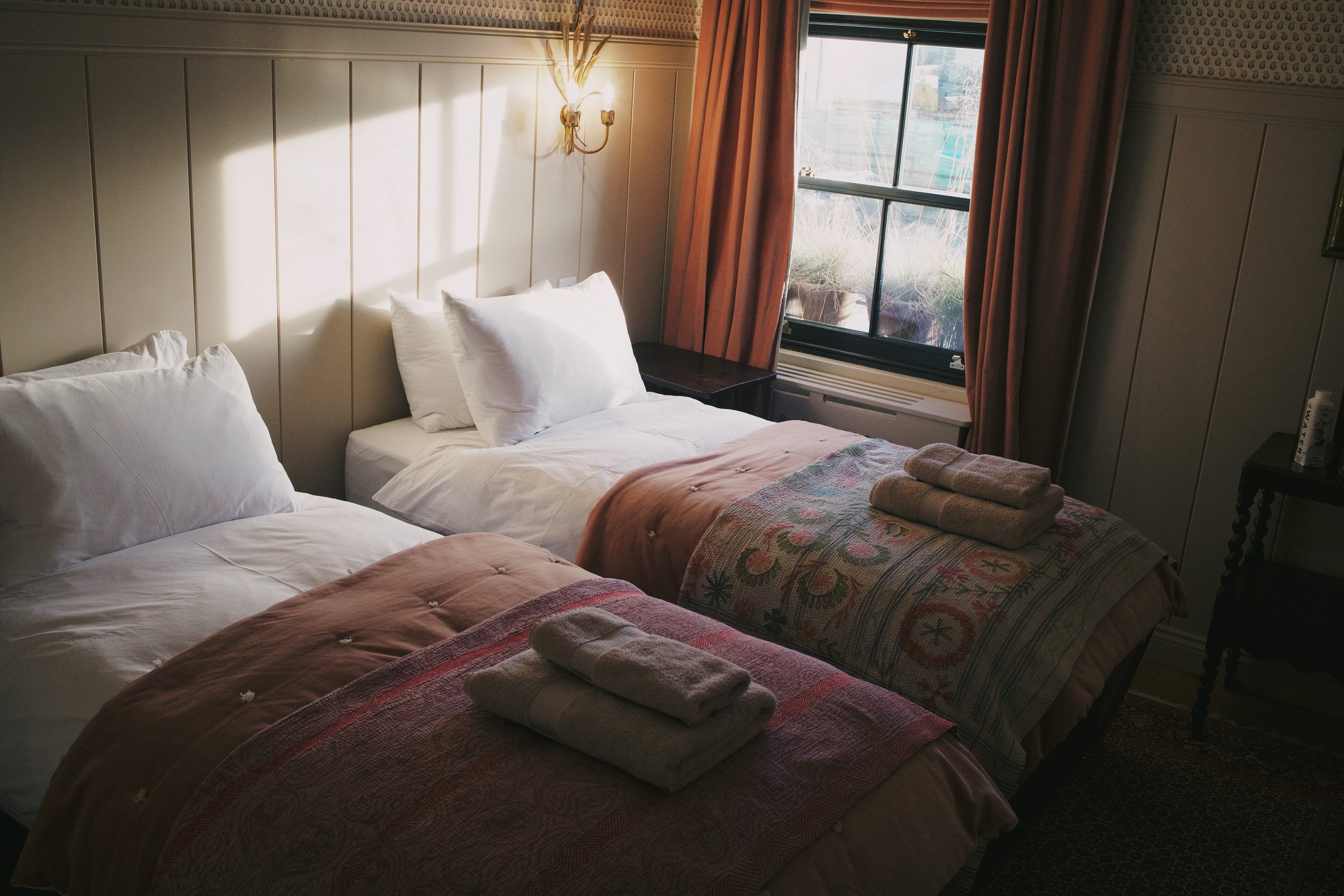 Two single beds with white bedding and pink patterned blankets, folded towels on top, in a warmly lit hotel room with a window, curtains, and wall-mounted lights.