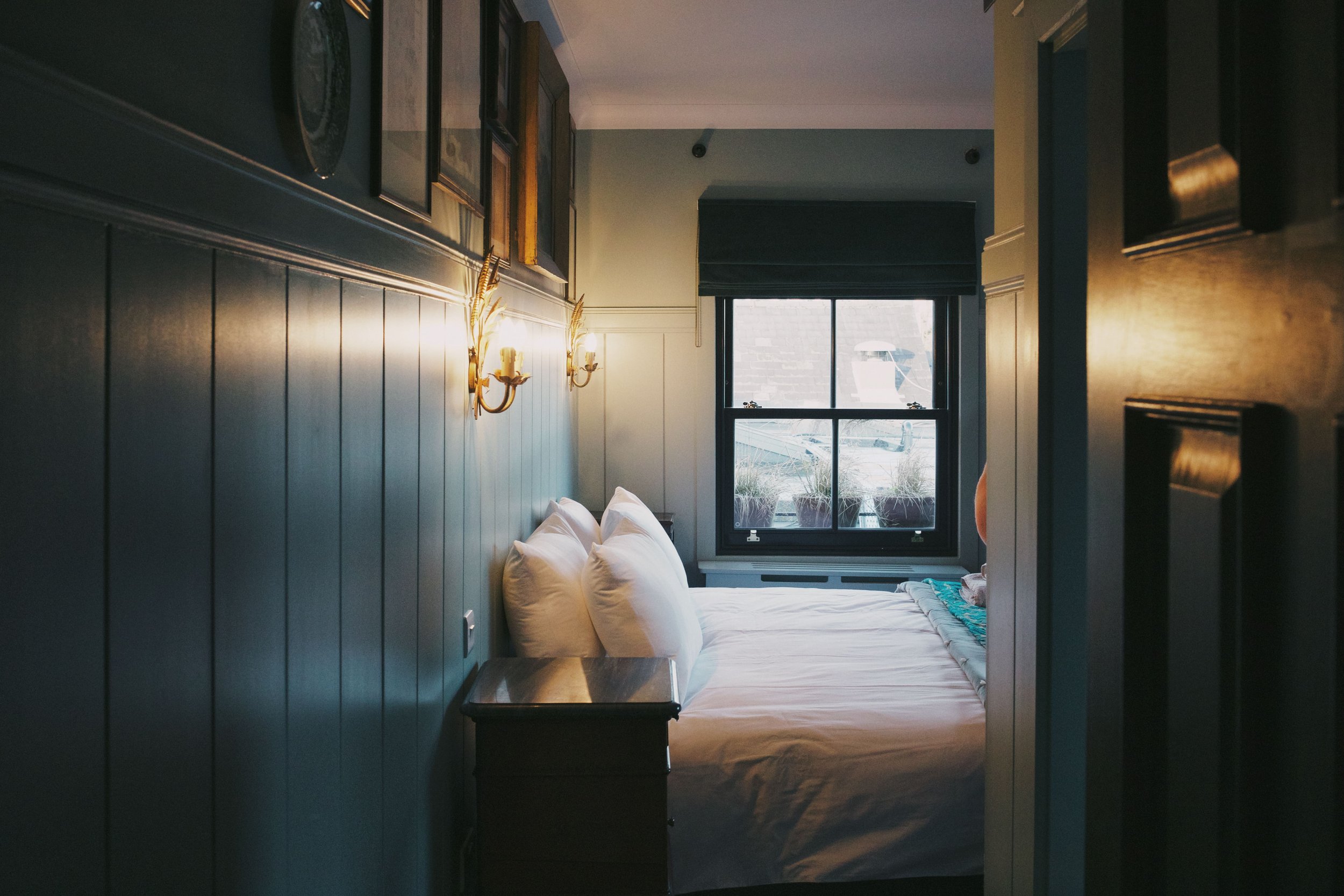 A cozy bedroom with a bed, white pillows, and a window showing boats outside, illuminated by wall sconces and natural light.