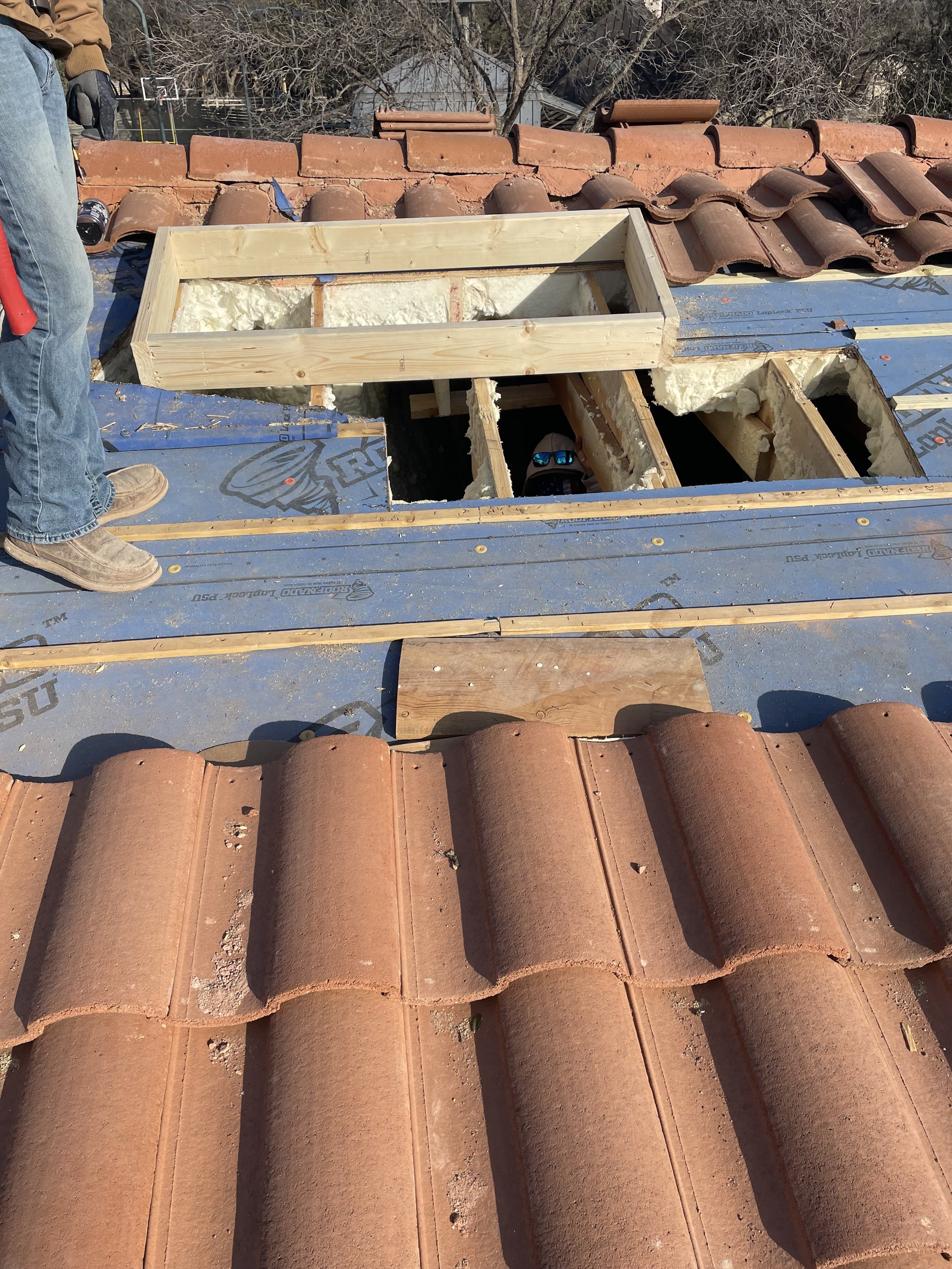 Roof construction site with clay tiles and a worker standing beside an open section of the roof, showing wooden framing and insulation.